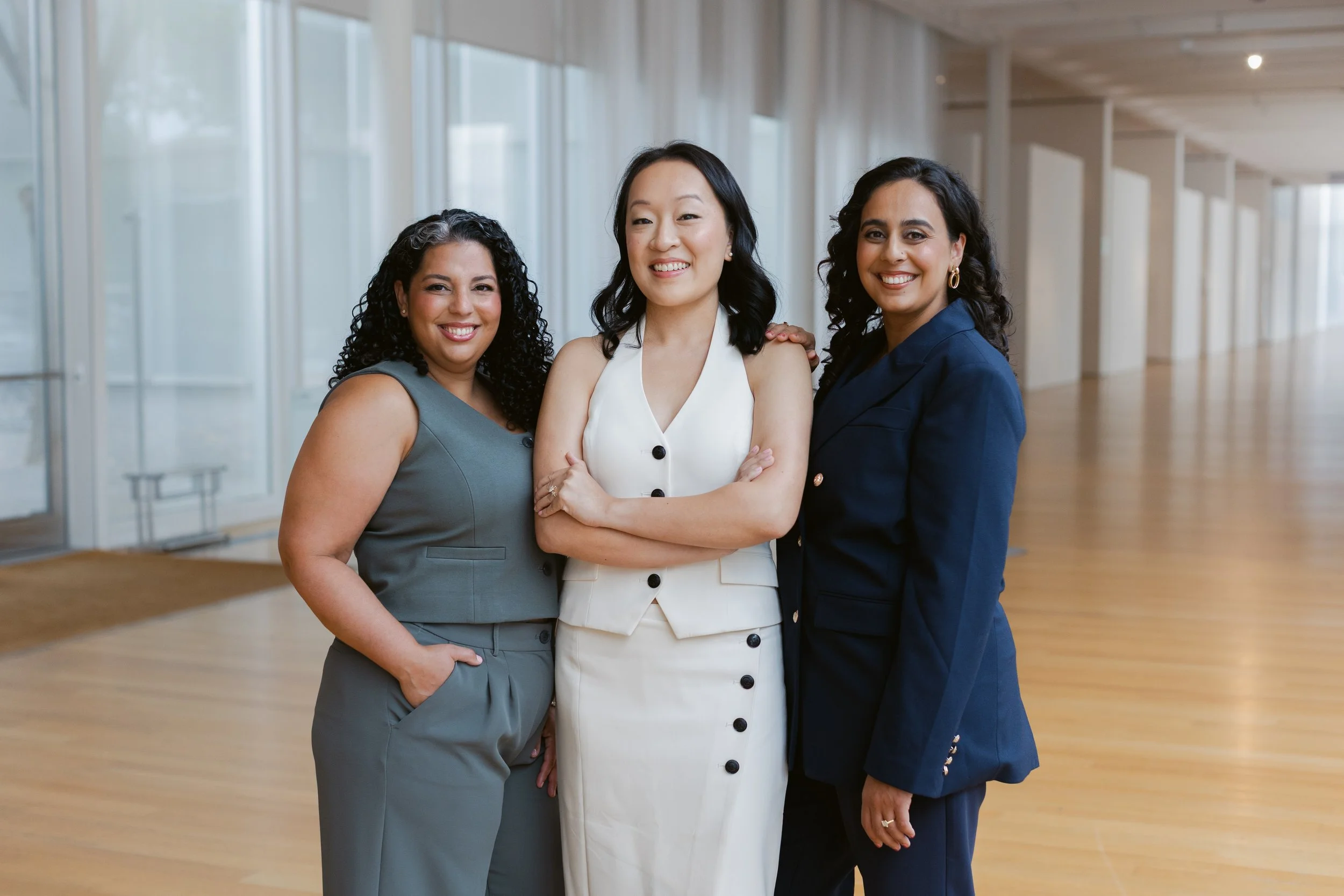 Three women standing together in a modern, bright interior, smiling at the camera.