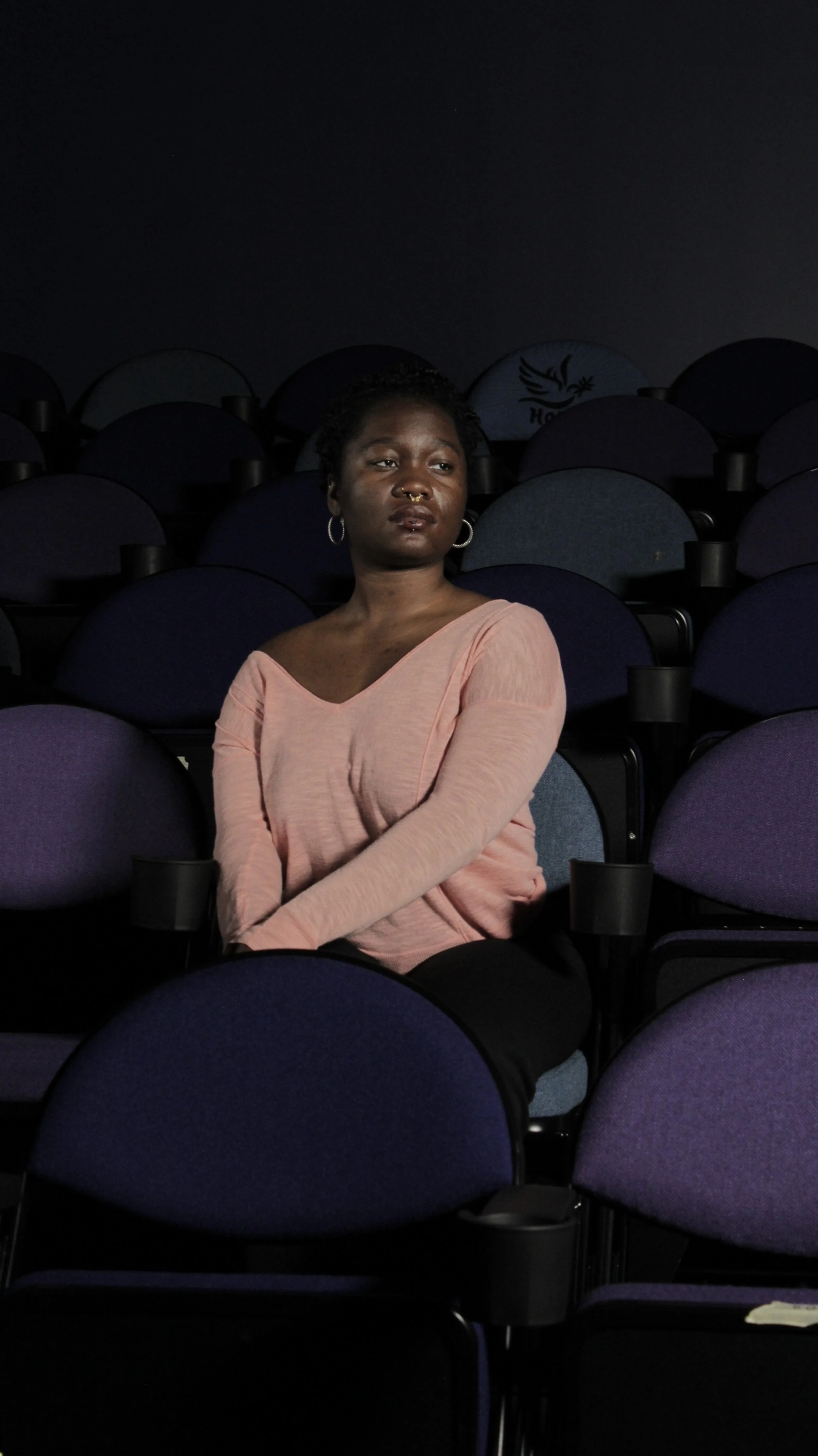 A young woman with dark skin, short curly hair, wearing hoop earrings, a nose ring, and a light pink off-shoulder top, sitting alone in a dark auditorium with mostly empty purple seats around her.
