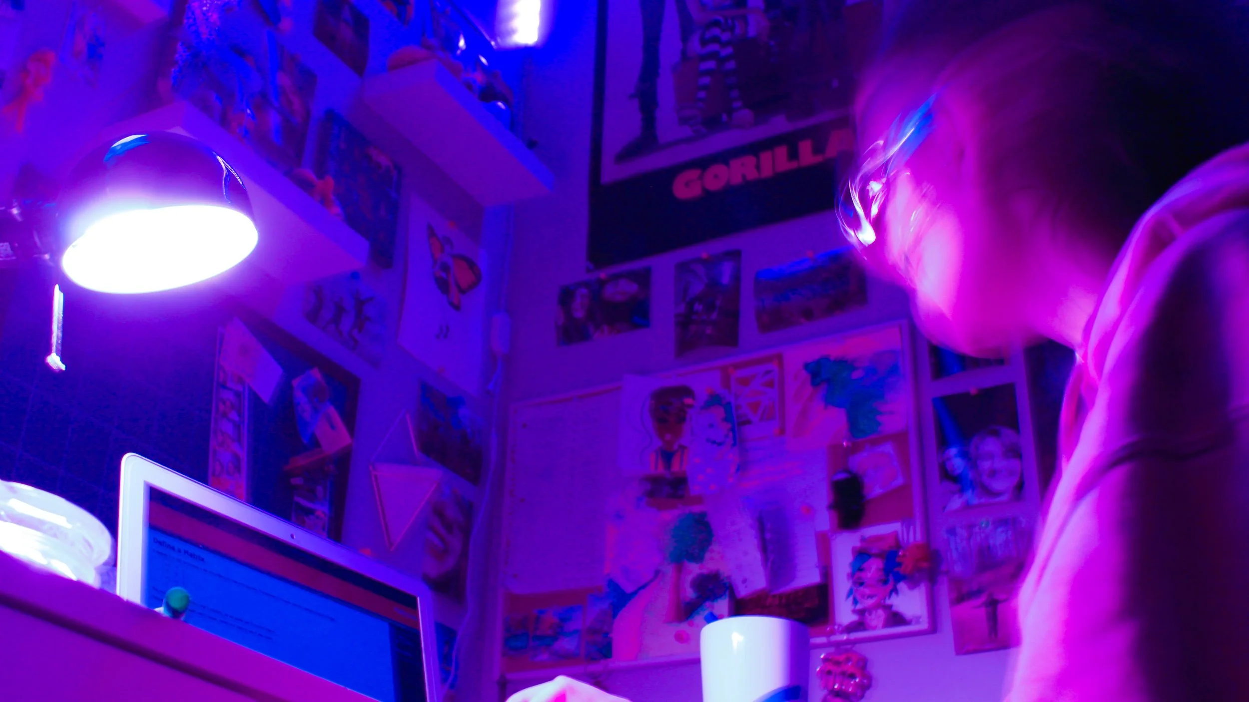 Person sitting in a room illuminated by purple light, with a wall covered in pictures, posters, and artwork, and a desk with a laptop.
