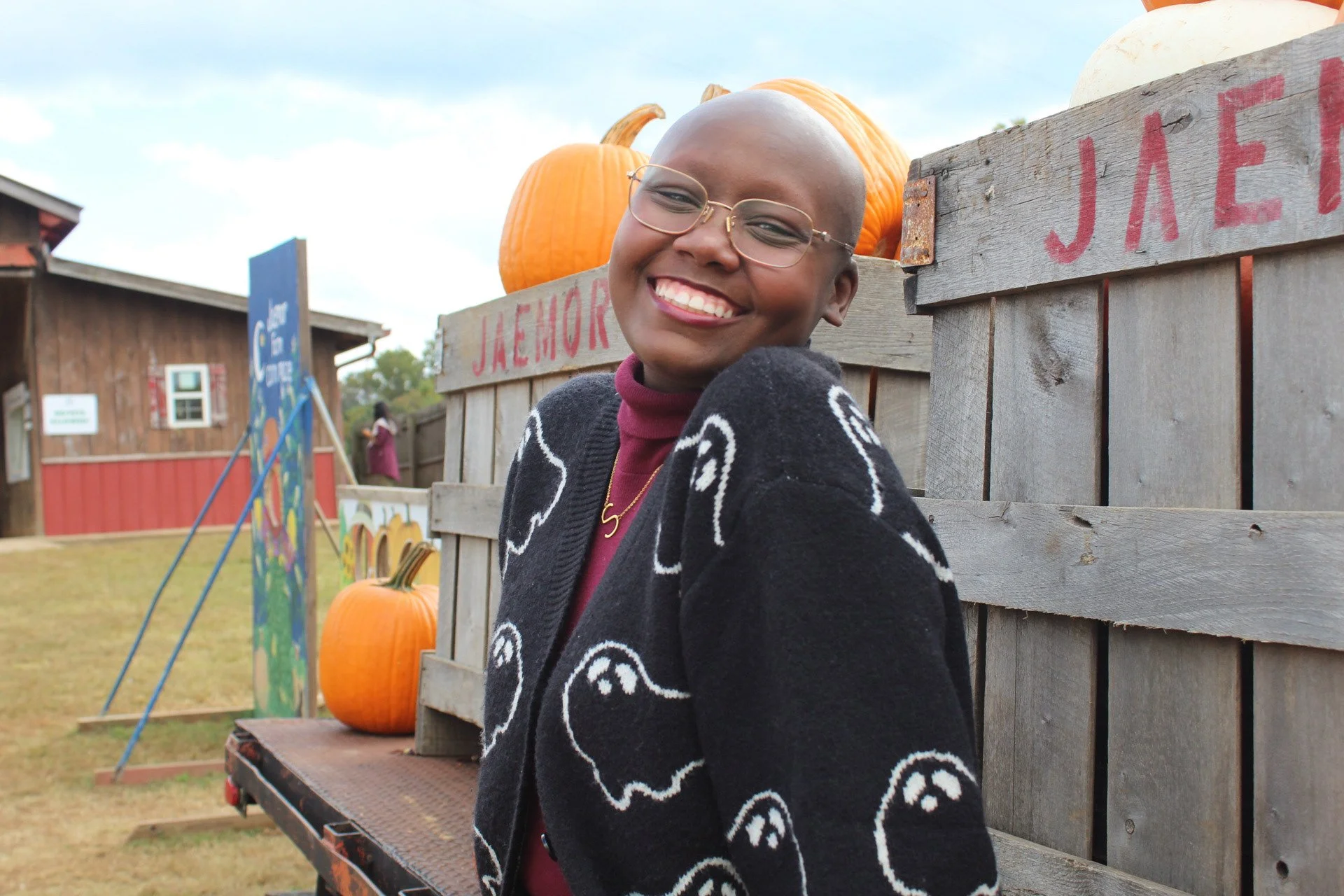 A smiling person with glasses and a black sweater with white ghost patterns at a pumpkin patch.