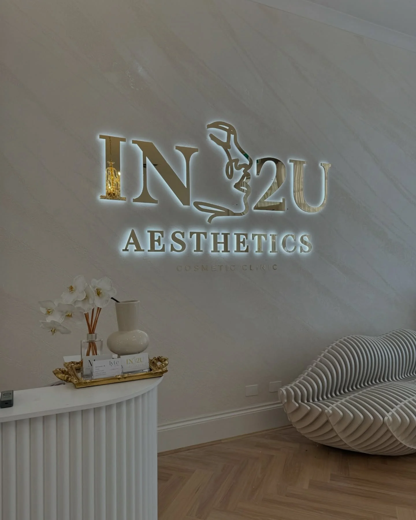Interior of a cosmetic clinic with illuminated sign that reads 'IN2U Aesthetics' and a decorative remote on a white, textured reception desk