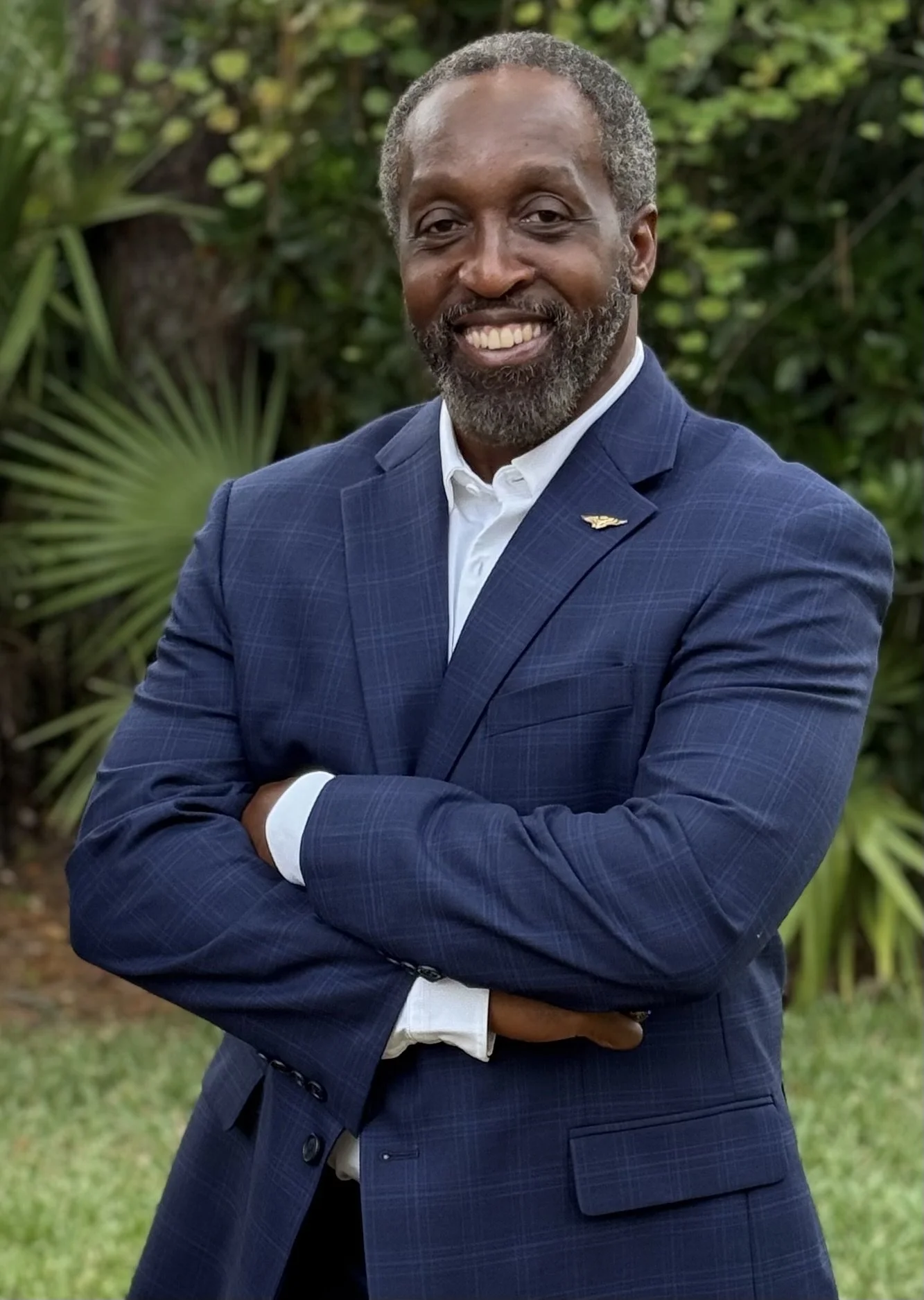 A smiling Black professional executive in a blue checked suit with his arms crossed, standing outdoors with greenery in the background.