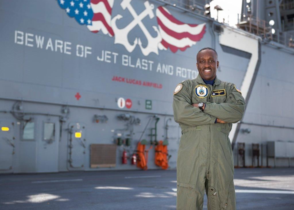 A smiling African American naval officer in uniform standing on the deck of an aircraft carrier, with a large Navy insignia and American flag graphics behind him.