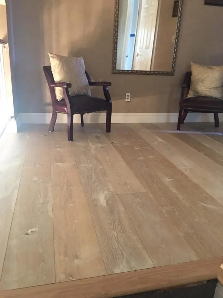 A room with wooden flooring, two dark wood chairs with beige cushions, a mirror on the wall, and a door reflected in the mirror.