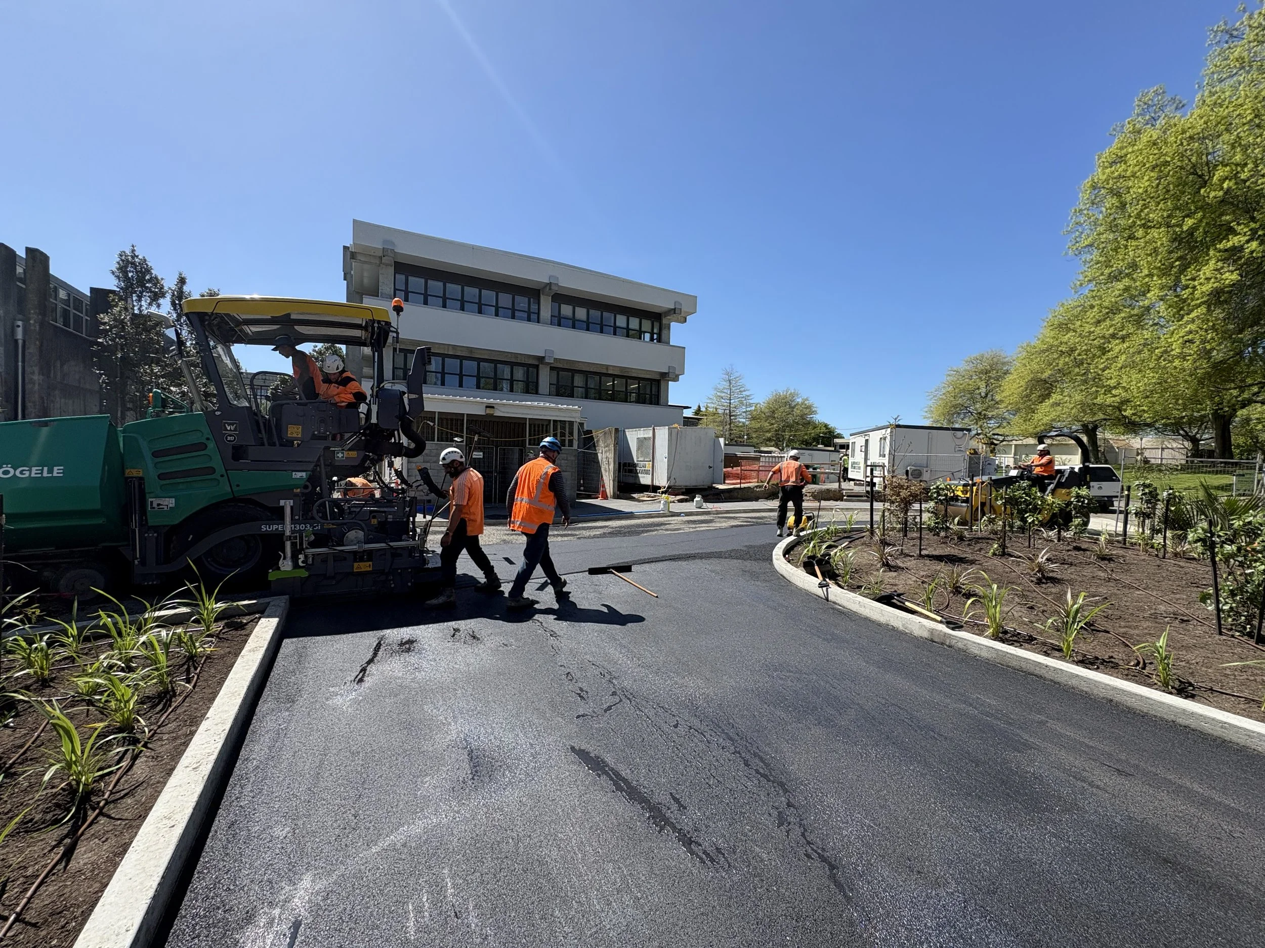 Construction workers paving a curved road in front of a modern building on a sunny day, with trees and landscaping to the side.