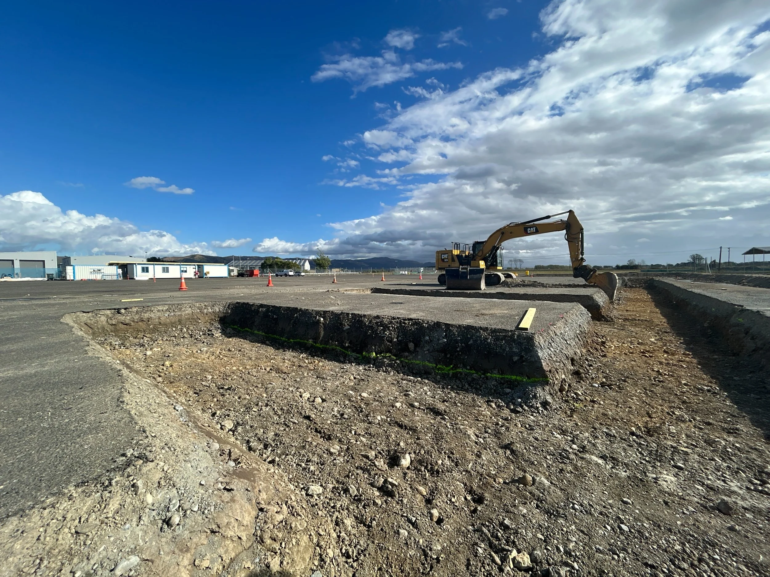 Construction site with a yellow excavator digging a trench under a partly cloudy sky.