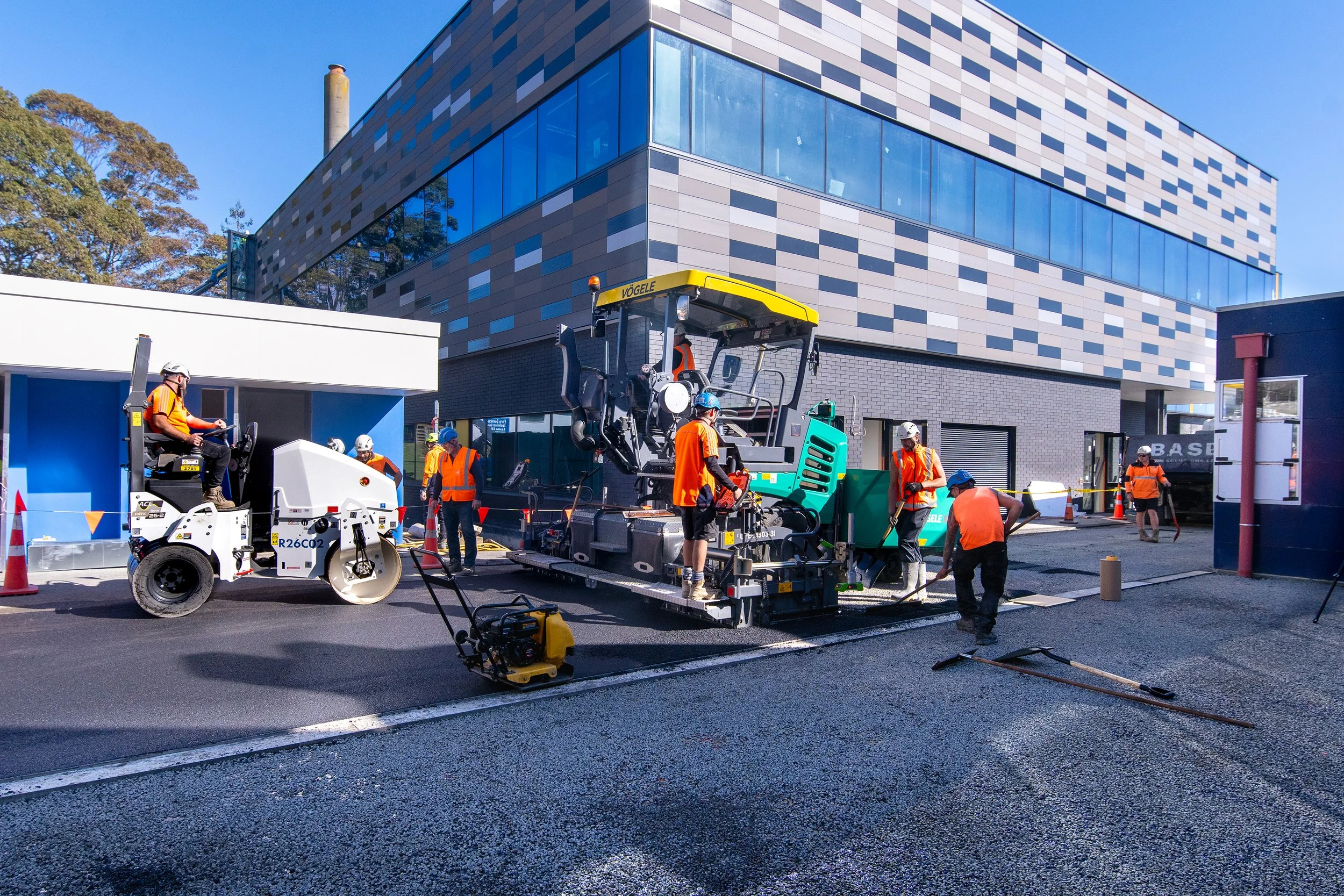Construction workers paving a road in front of a modern building with large glass windows, wearing orange safety vests and helmets, using heavy machinery and equipment.