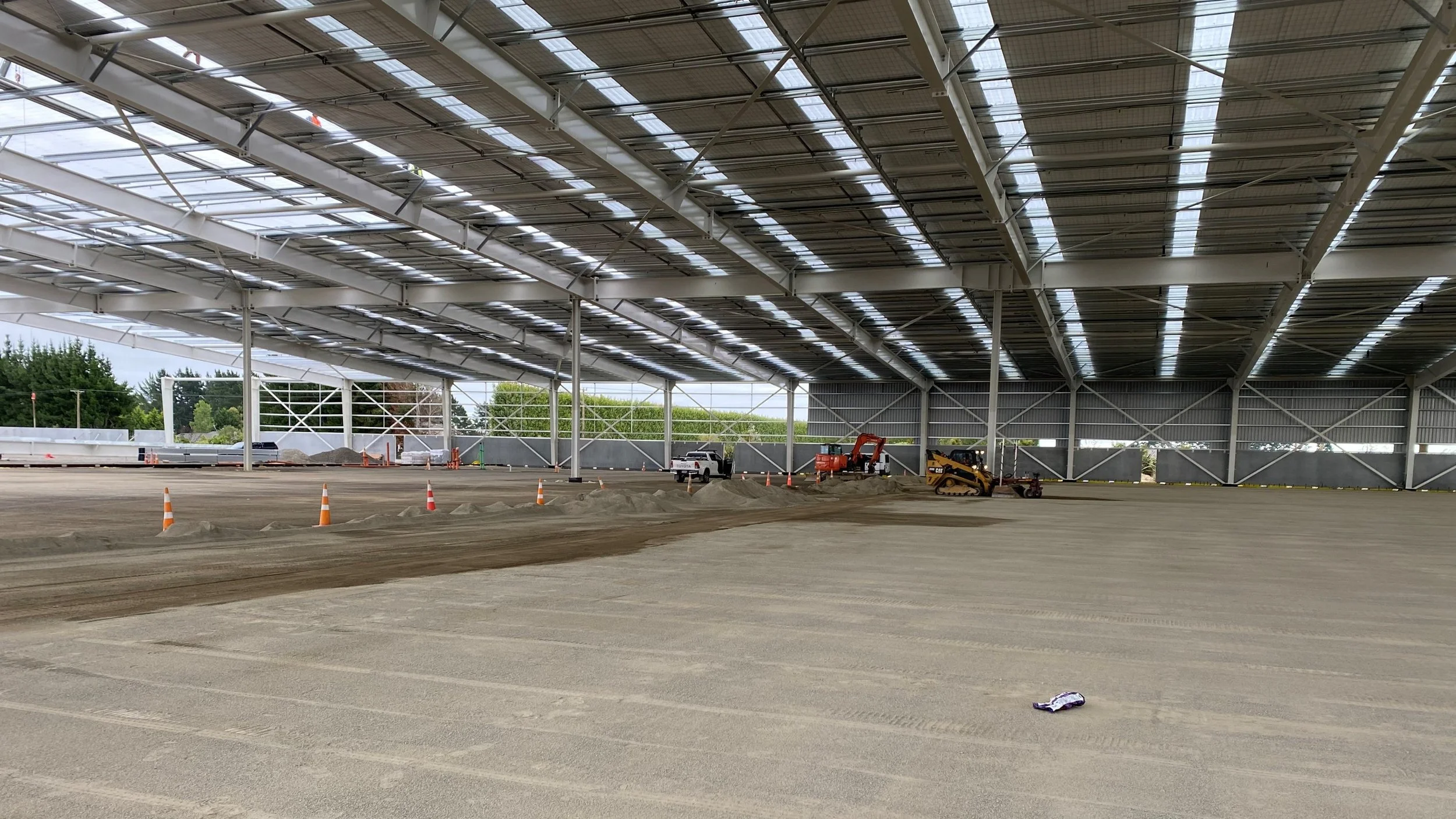 Inside a large warehouse or hangar under construction with solar panels installed on the roof. There are construction equipment, including a small vehicle, a mini excavator, and a roller, working on the ground. Orange cones line the area, and there i