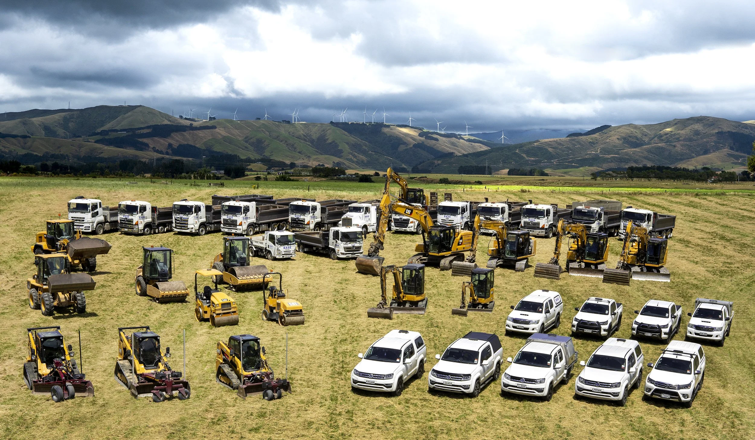 A large collection of construction and utility vehicles on a grassy field with hills and wind turbines in the background.