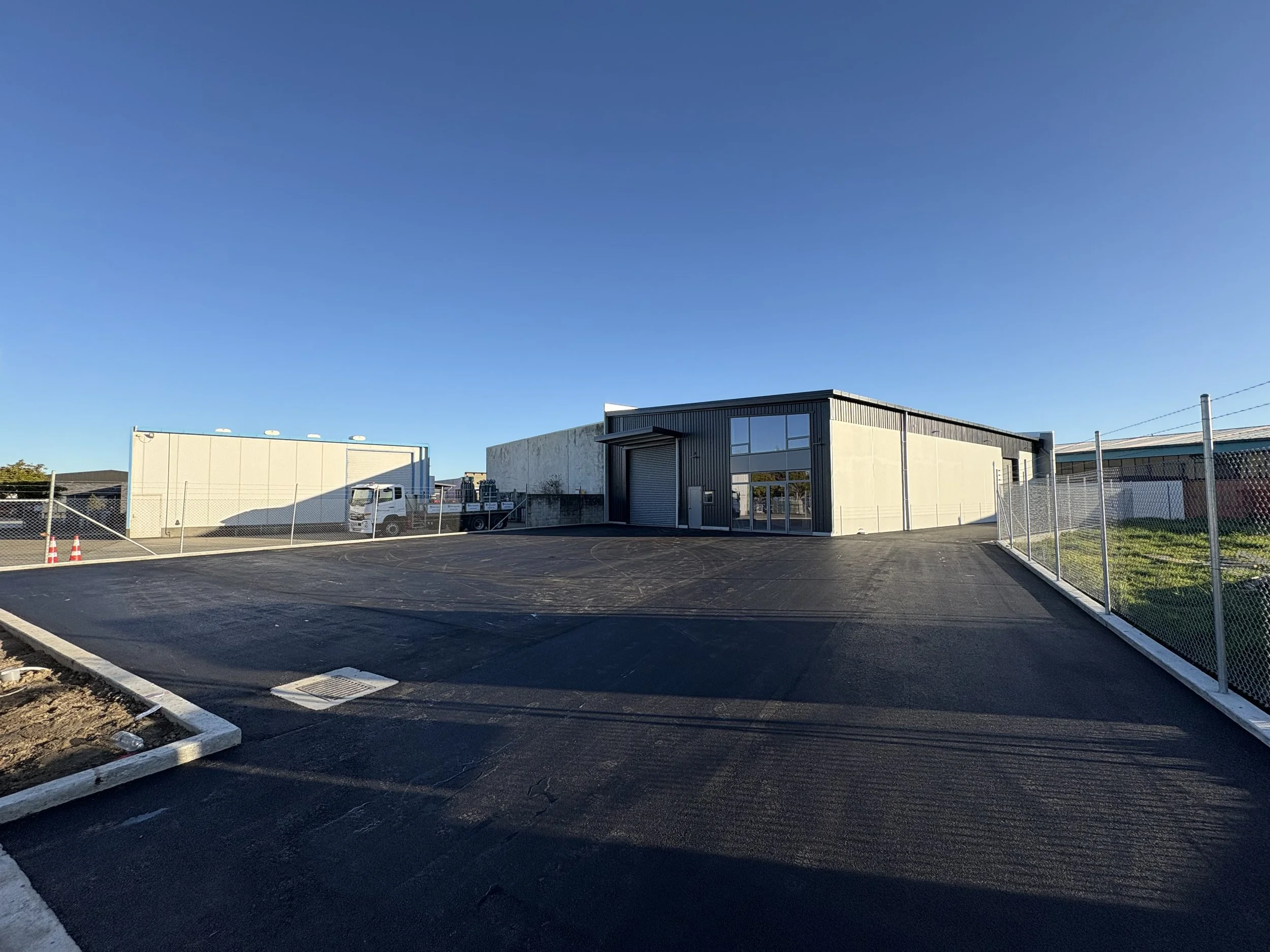 Empty black asphalt parking lot with a newly paved surface, fenced on two sides, and a modern industrial building with large windows and a roller door under a clear blue sky.