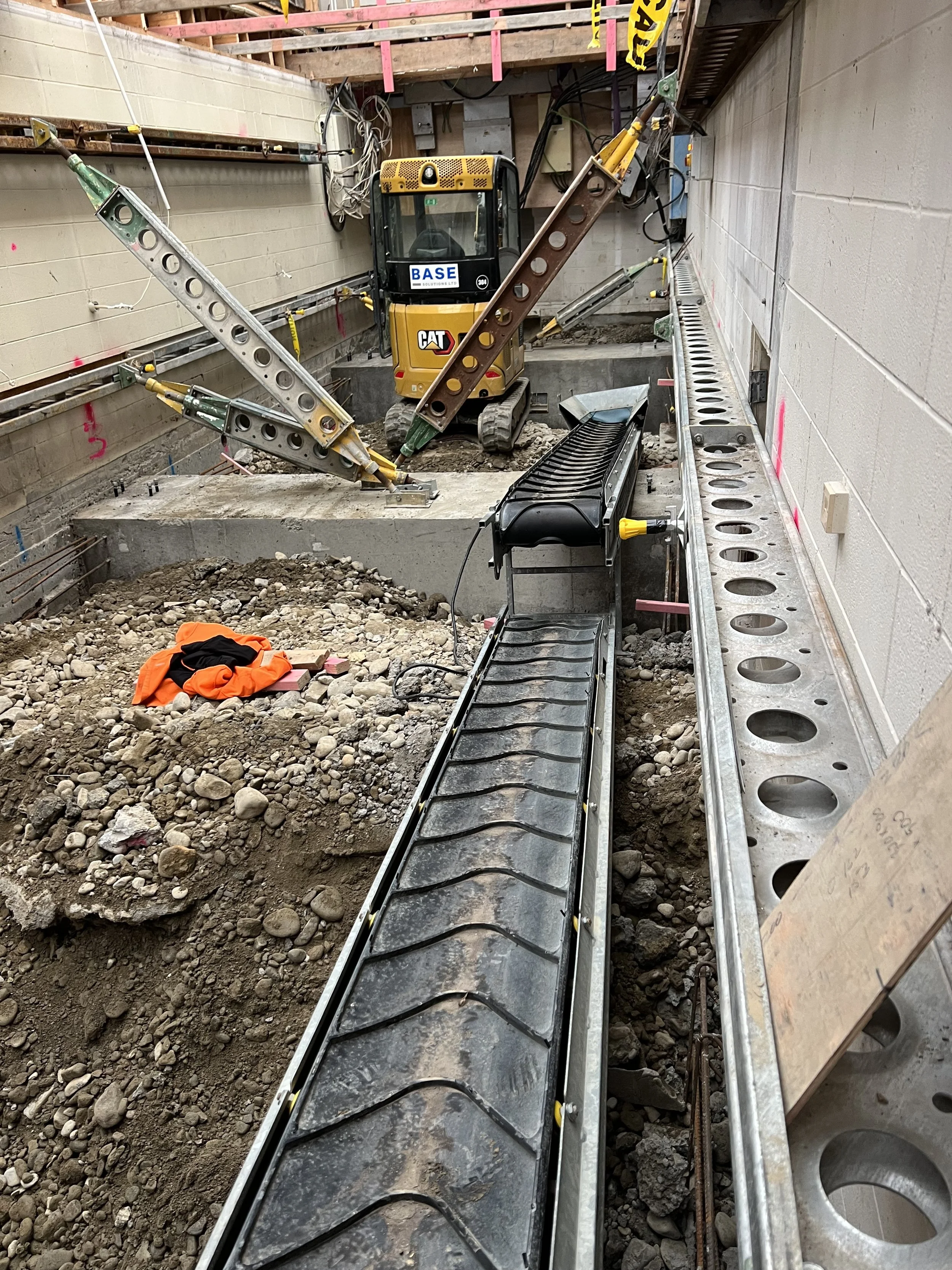 Construction site with equipment, a small excavator, steel beams, and a conveyor or drainage system in an indoor space.