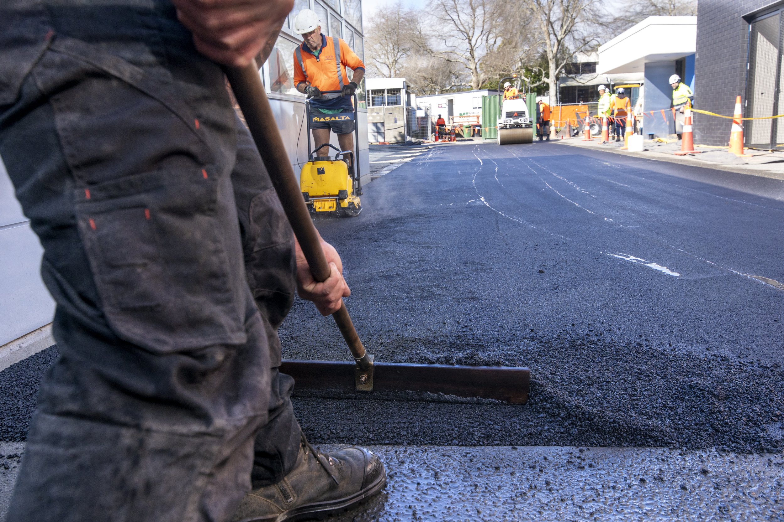 Close-up of construction worker's legs and hands using a rake tool to spread fresh asphalt on a road during daytime, with other construction workers and equipment visible in the background.