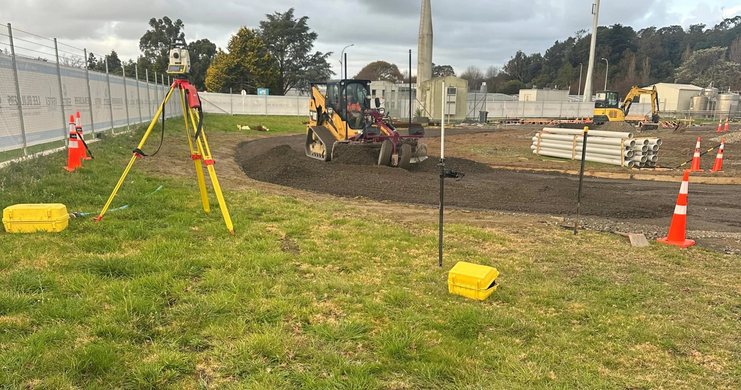 Construction site with machinery, orange cones, and pipes, including a bulldozer and a small excavator, on a cloudy day with a grassy area in the foreground.