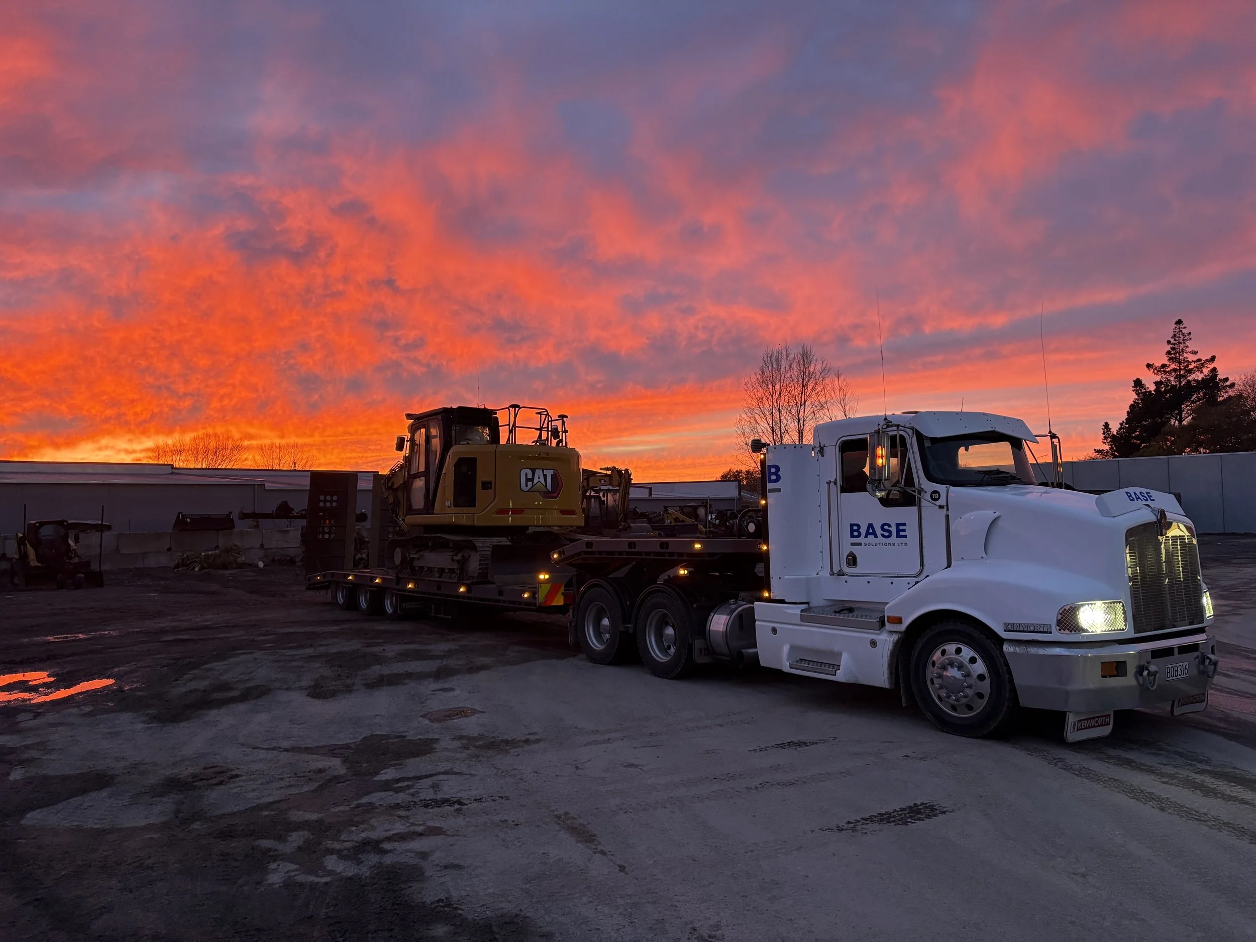 White semi-truck with company name BASE on the door, transporting a yellow CAT construction excavator at sunset, with a colorful sky and a few trees in the background.
