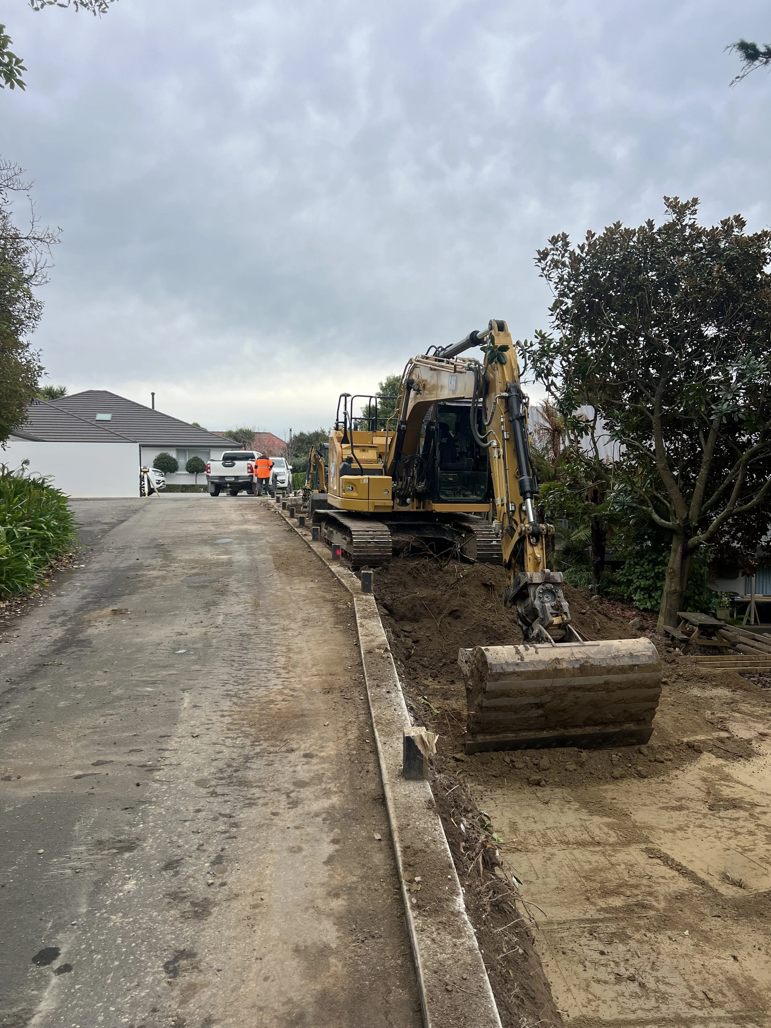Construction site with a backhoe loader digging along a prepared road area, with workers and parked cars visible in the background under an overcast sky.