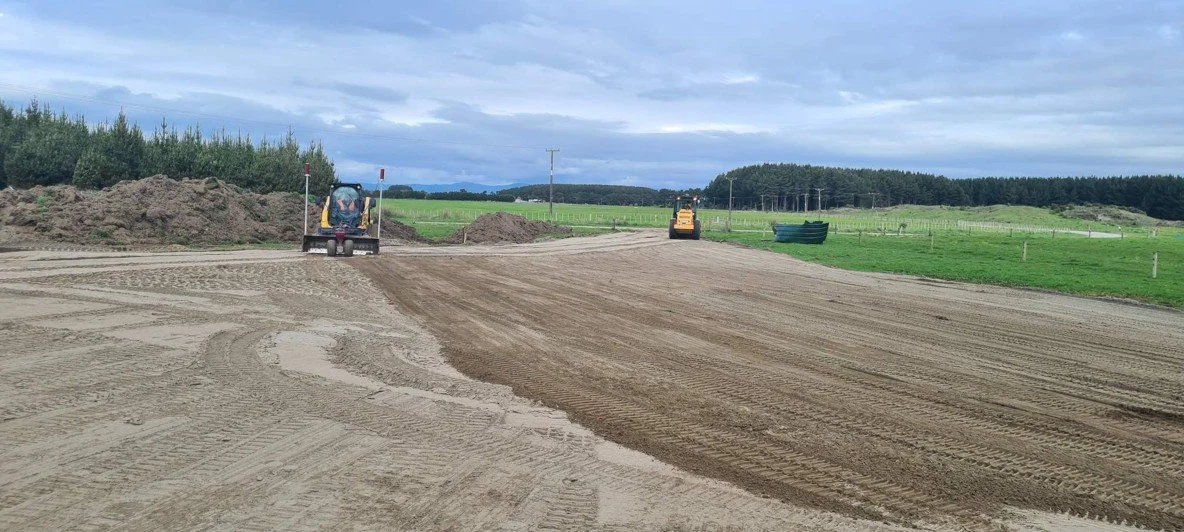 Construction site with dirt and gravel being leveled with bulldozers and compactors, rural landscape with trees and grassy fields in the background.