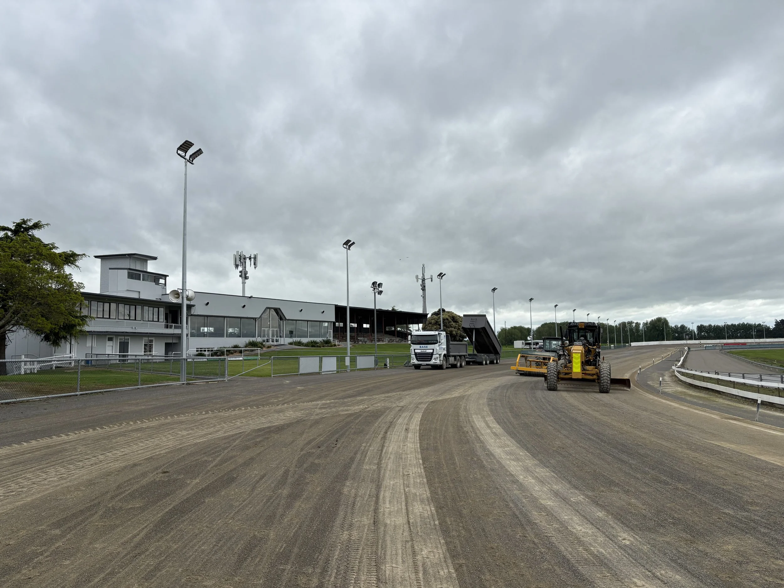 View of a horse racing track with construction equipment, including a grader and dump truck, working on the dirt infield. There's a modern building with large windows and floodlights on tall poles under a cloudy sky.
