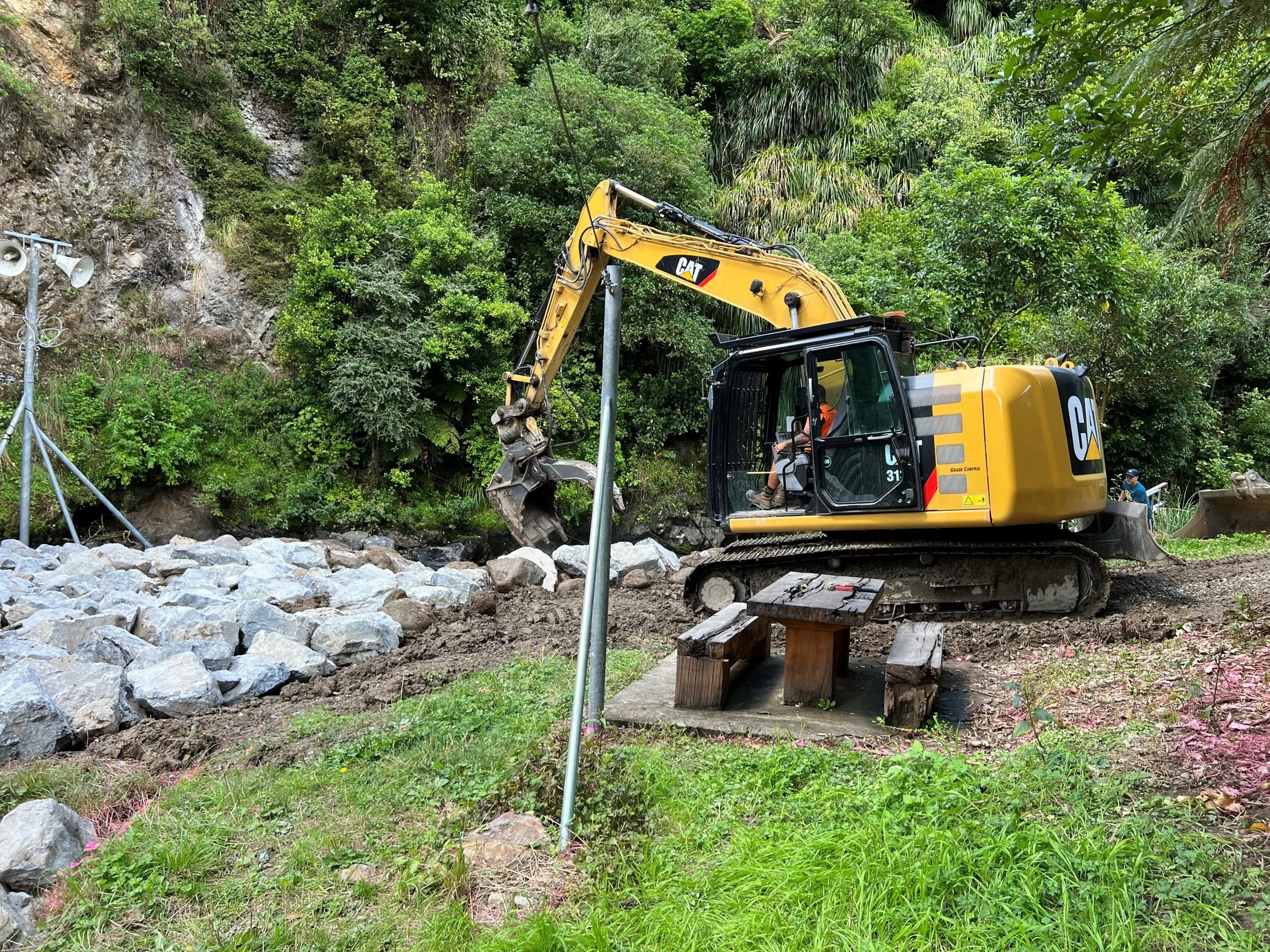 A yellow CAT excavator working on a rocky area outdoors with green trees and plants in the background. There are a couple of wooden benches and a small floodlight structure nearby.