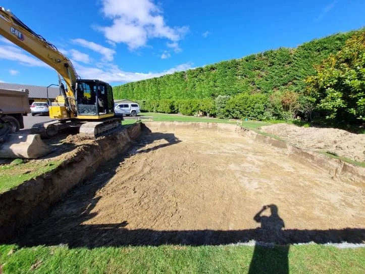 Construction site with a yellow excavator on dirt and a row of parked cars in the background under a clear blue sky.