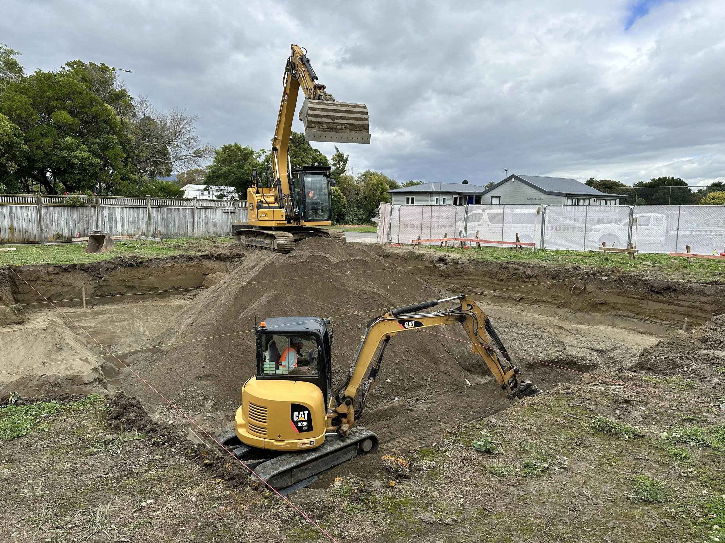 Two excavators working on a construction site, digging a large hole in the ground, with a cloudy sky overhead and residential buildings in the background.
