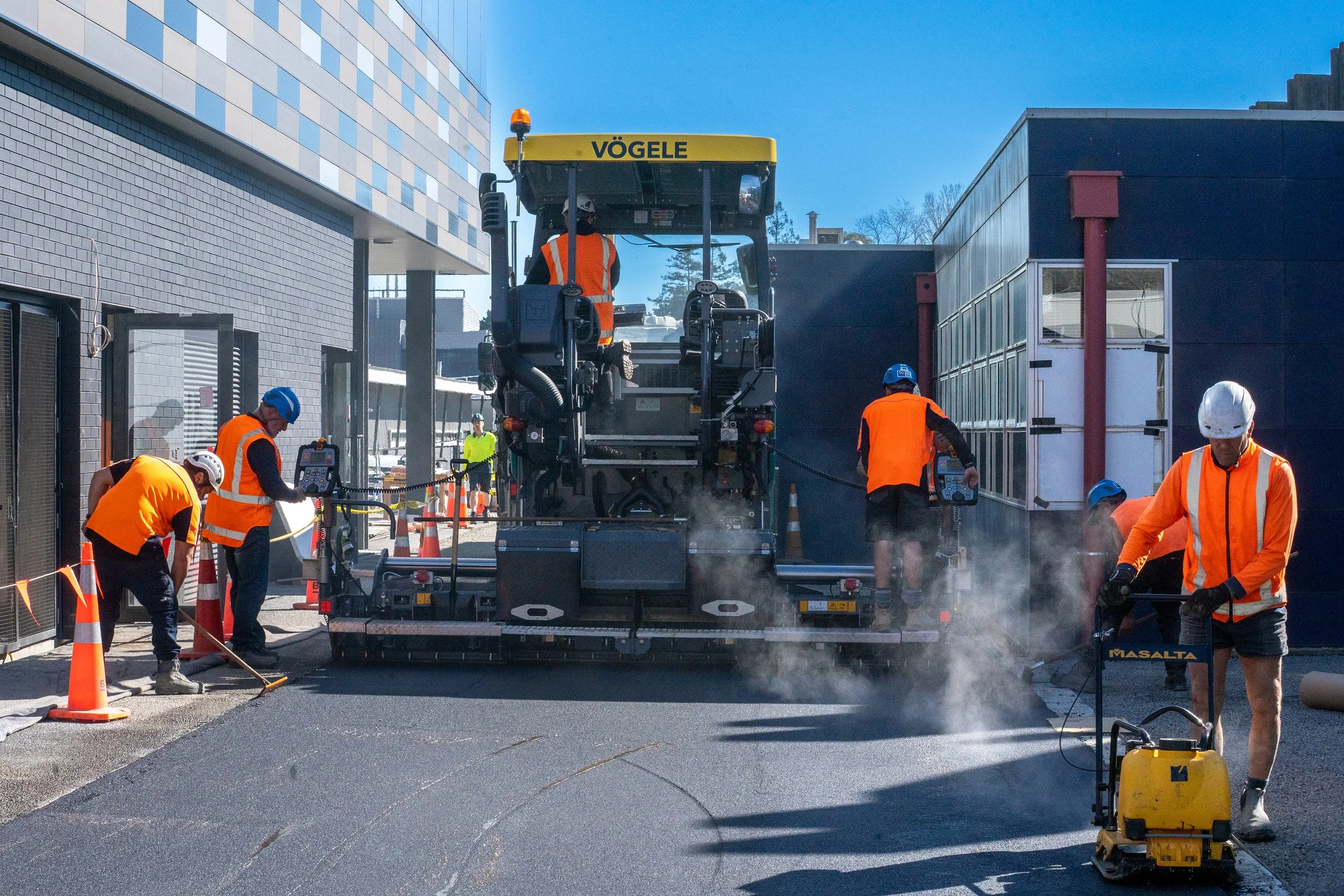 Workers in orange safety vests and helmets paving a road with a large asphalt paver machine branded "VÖGELE."