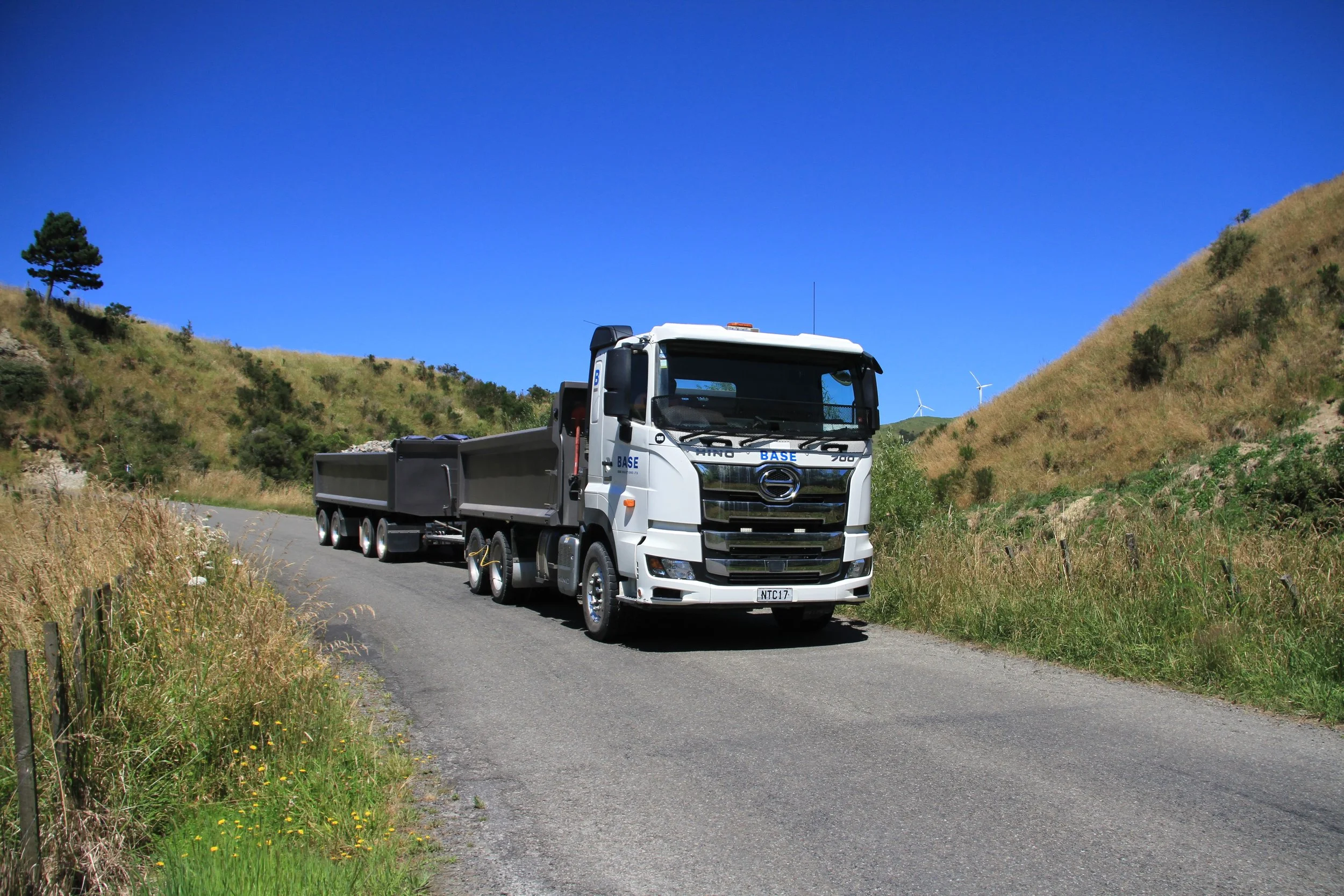 A white truck with a trailer driving on a rural mountain road with grassy hills and wind turbines in the background, under a clear blue sky.