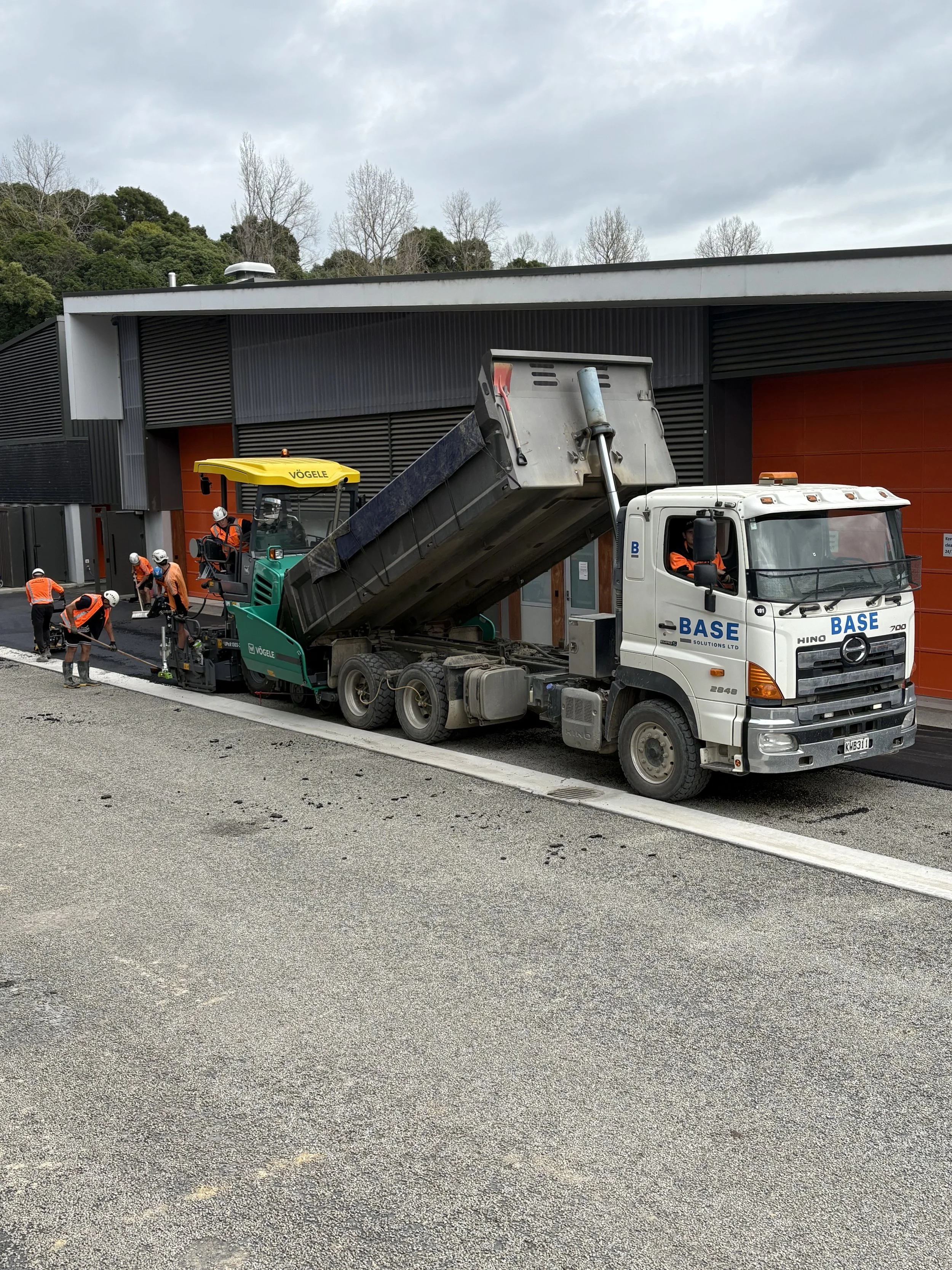 Construction workers paving a road next to a building, with a dump truck parked nearby.