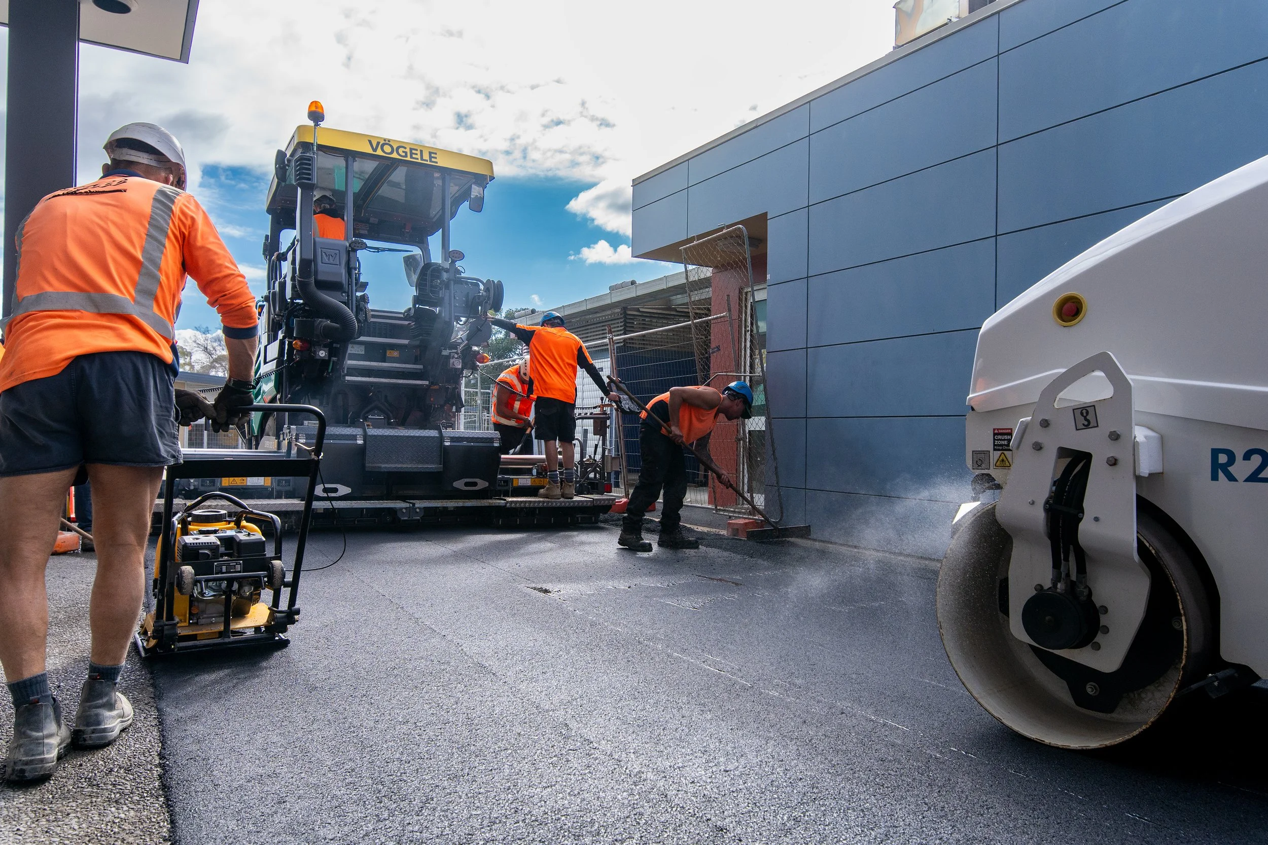 Construction workers and machines paving a road outdoors under a cloudy sky.
