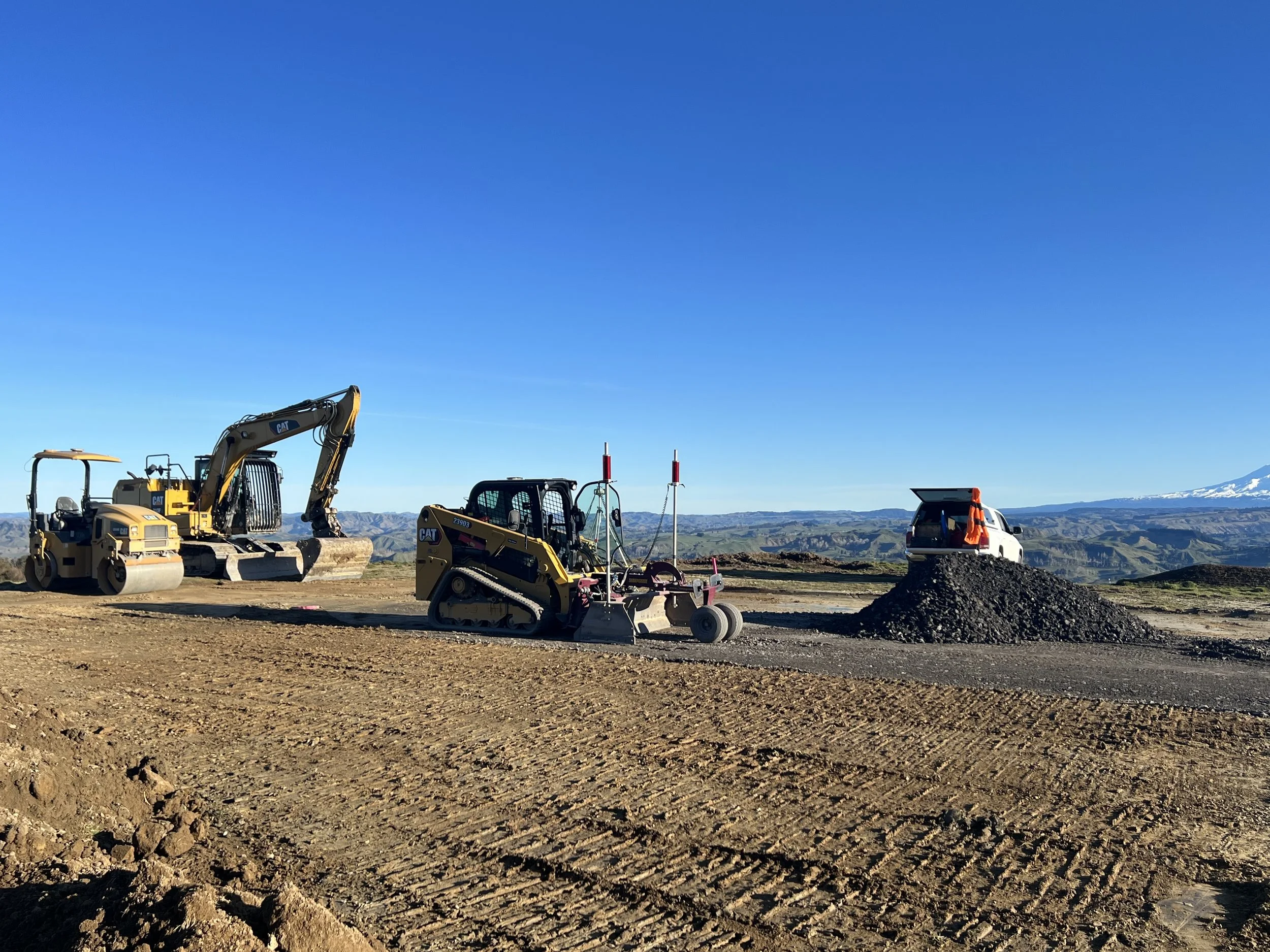 Construction site with bulldozer, roller, and a small truck, with mountains in the background and clear blue sky.