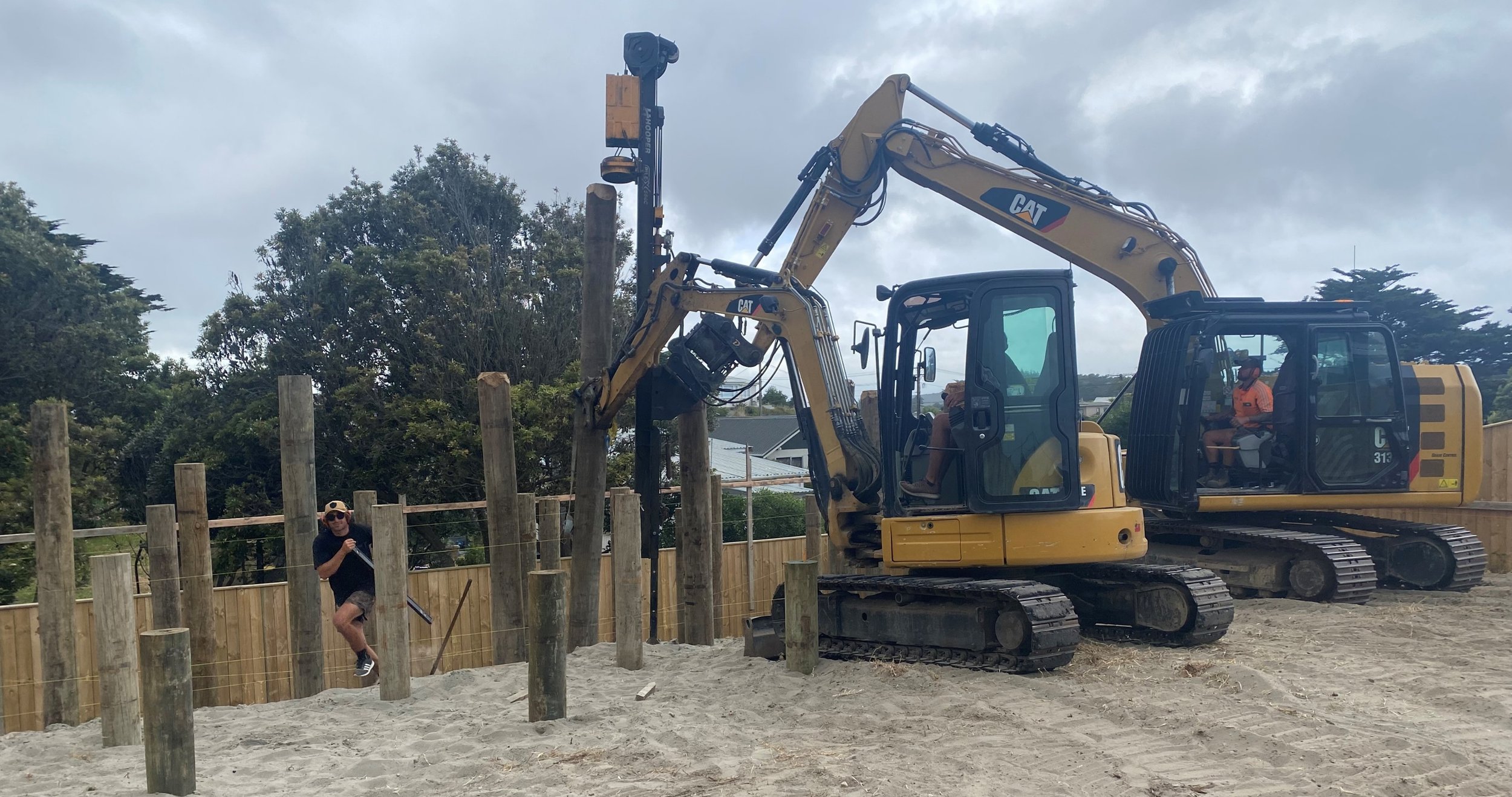 Two yellow excavators with black tracks working on construction, one with a man sitting inside, and another man in sunglasses and a black shirt climbing wooden posts nearby on a dirt surface with a wooden fence behind and trees in the background.