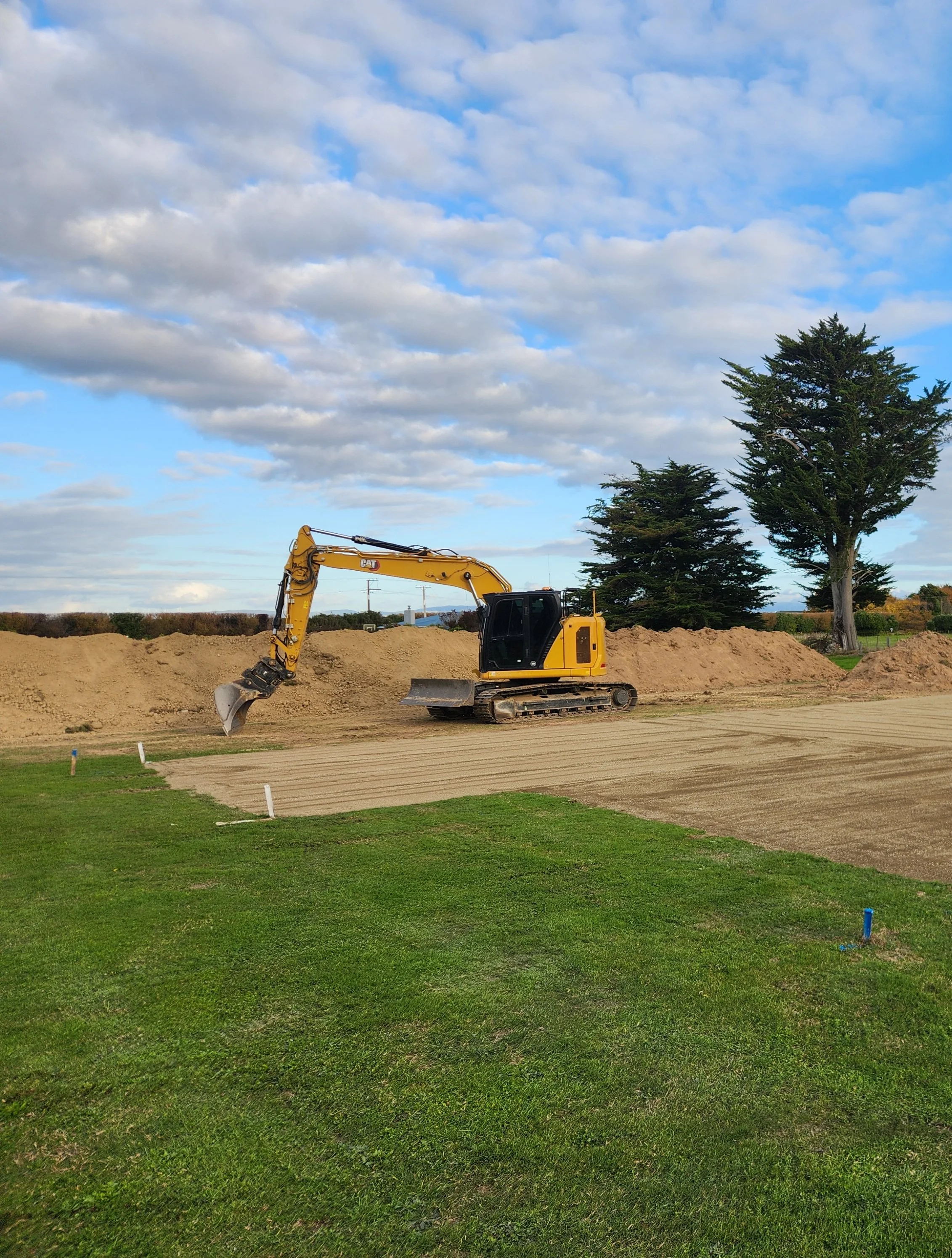 A yellow excavator working on a construction site, with mounds of dirt and trees in the background, under a partly cloudy sky.
