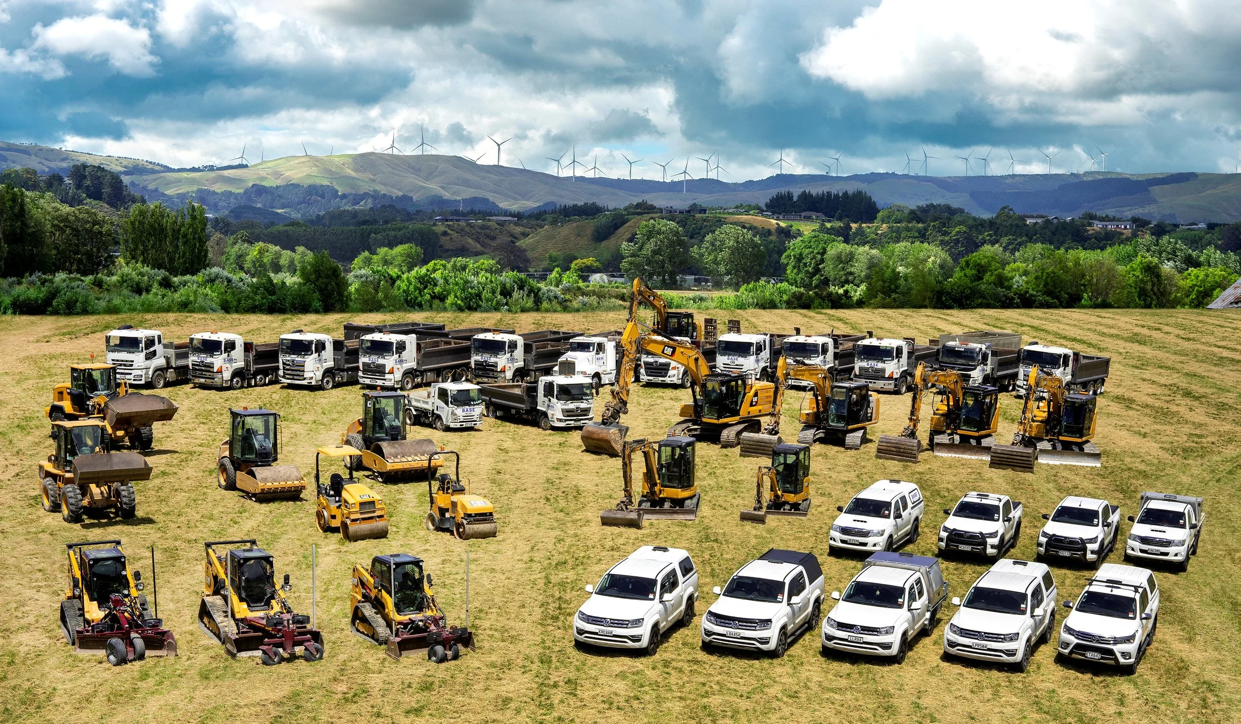 Construction and utility vehicles arranged on a grassy field with a backdrop of green hills and wind turbines under a cloudy sky.