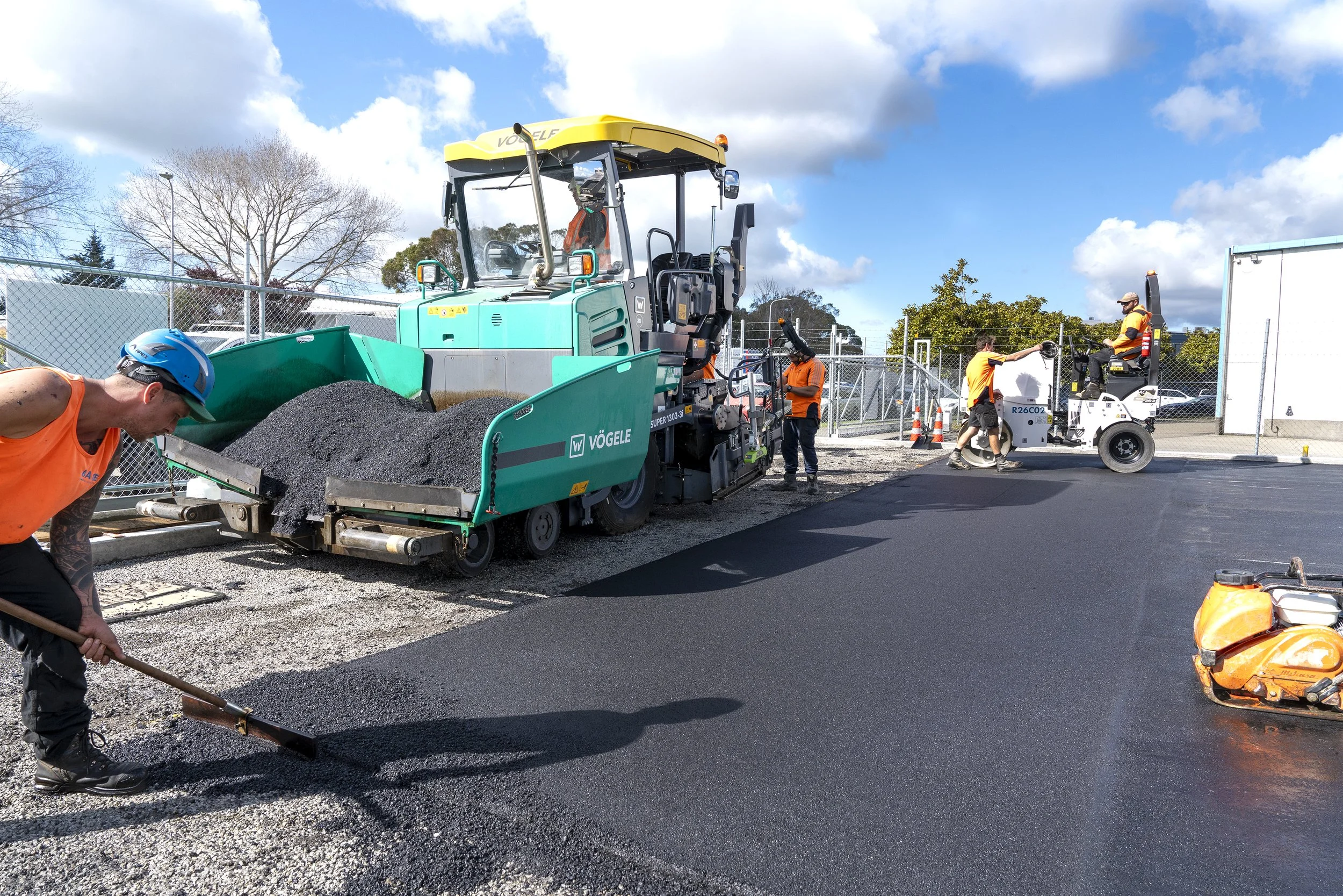 Construction workers paving a road with asphalt, using heavy machinery and tools on a sunny day.