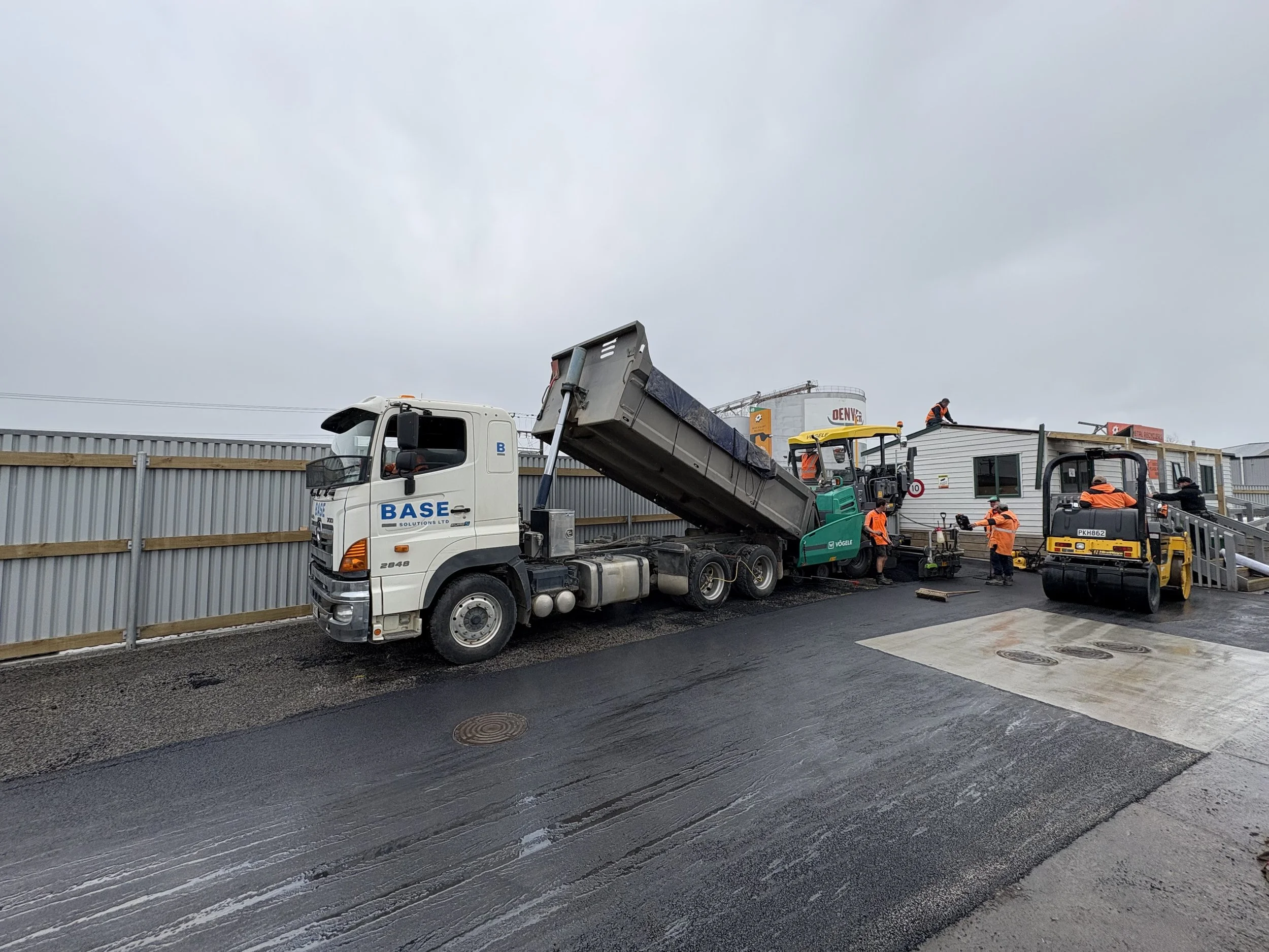Construction workers paving asphalt on a road next to a white building with a metal fence and cloudy sky.