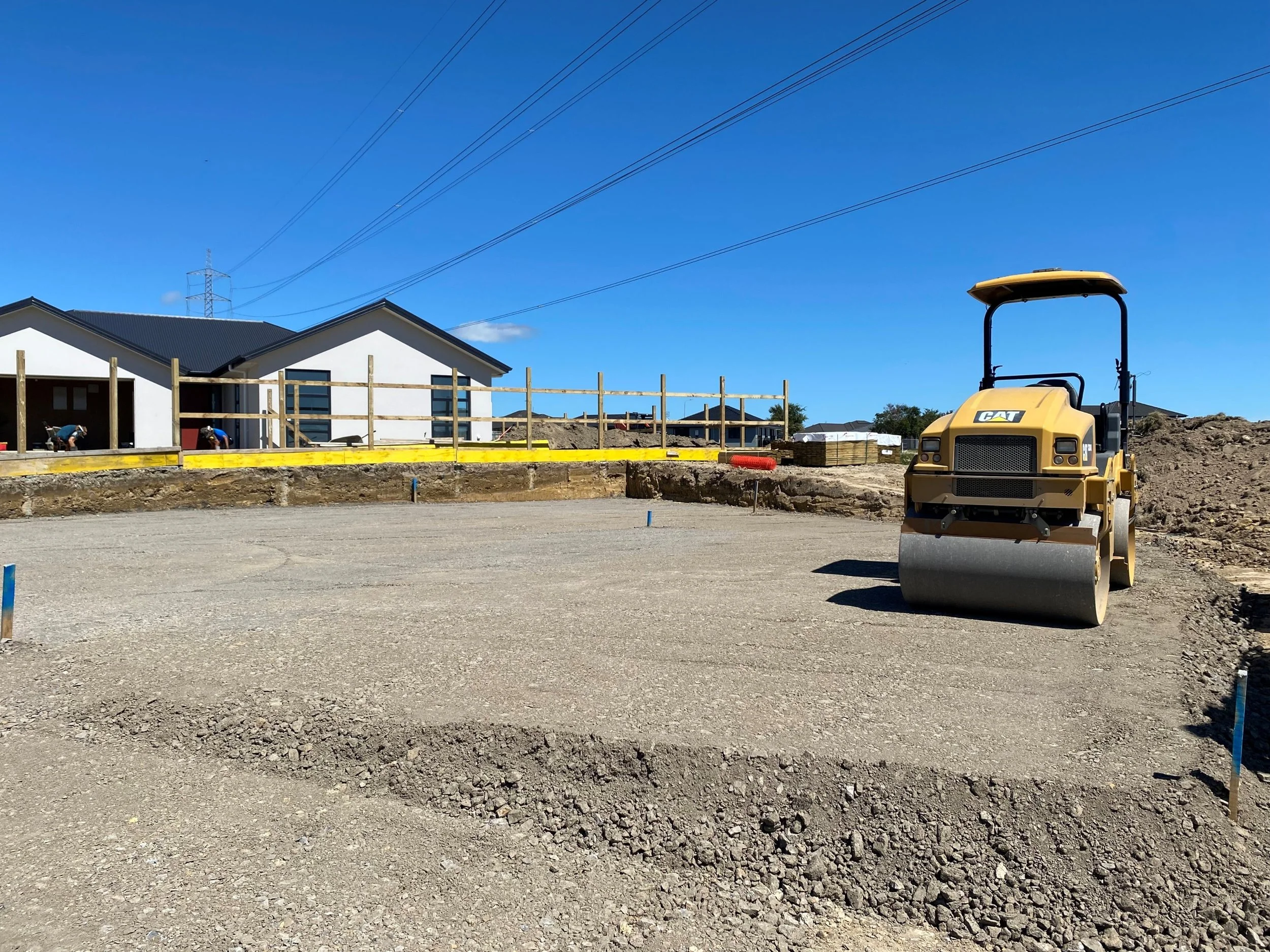 Construction site with a yellow steamroller, gravel, and ongoing building work in the background, under a blue sky.