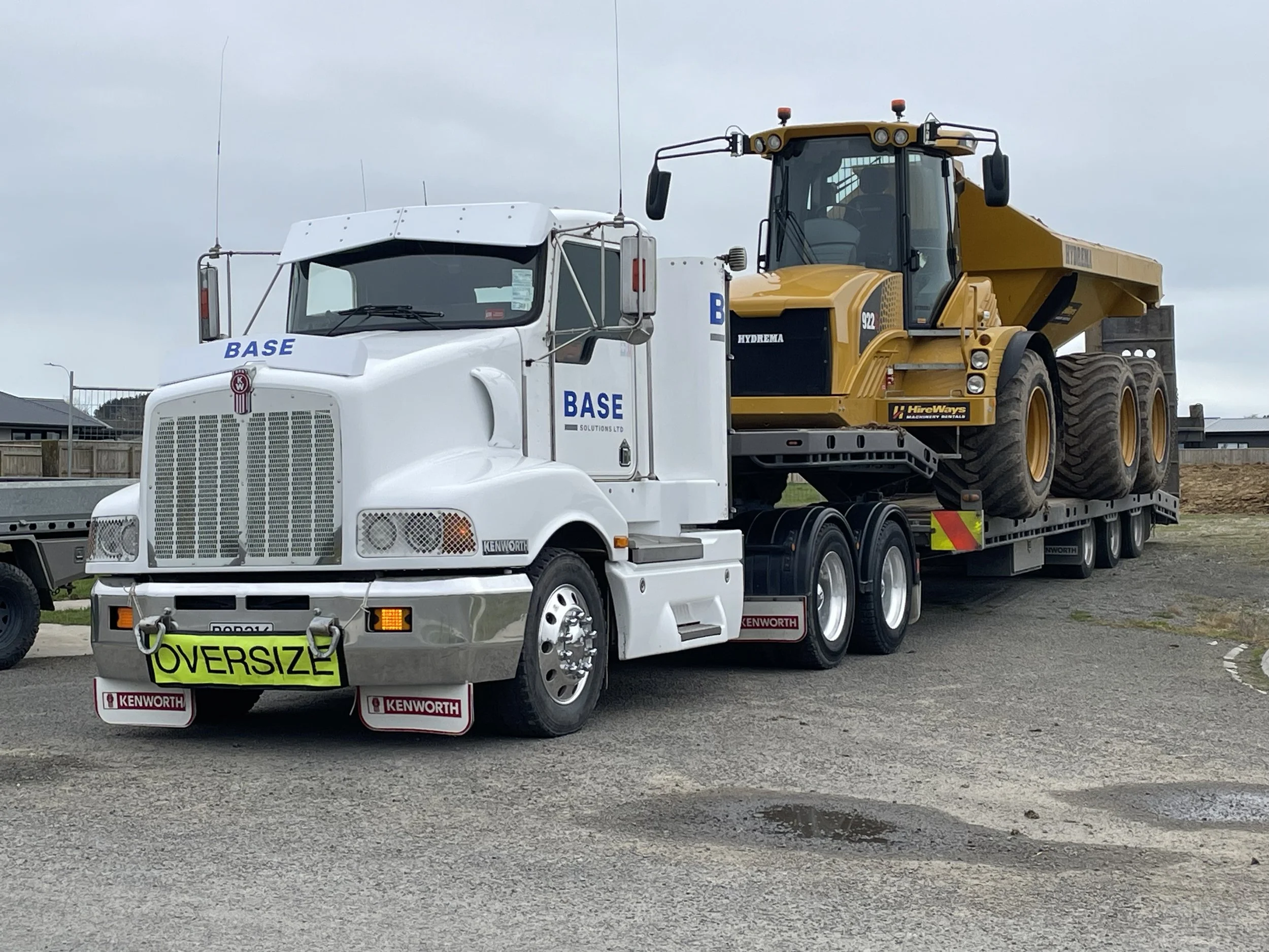 A white Kenworth semi-truck carrying a large yellow dump truck, with a bright yellow 'Oversize' sign on the front bumper.