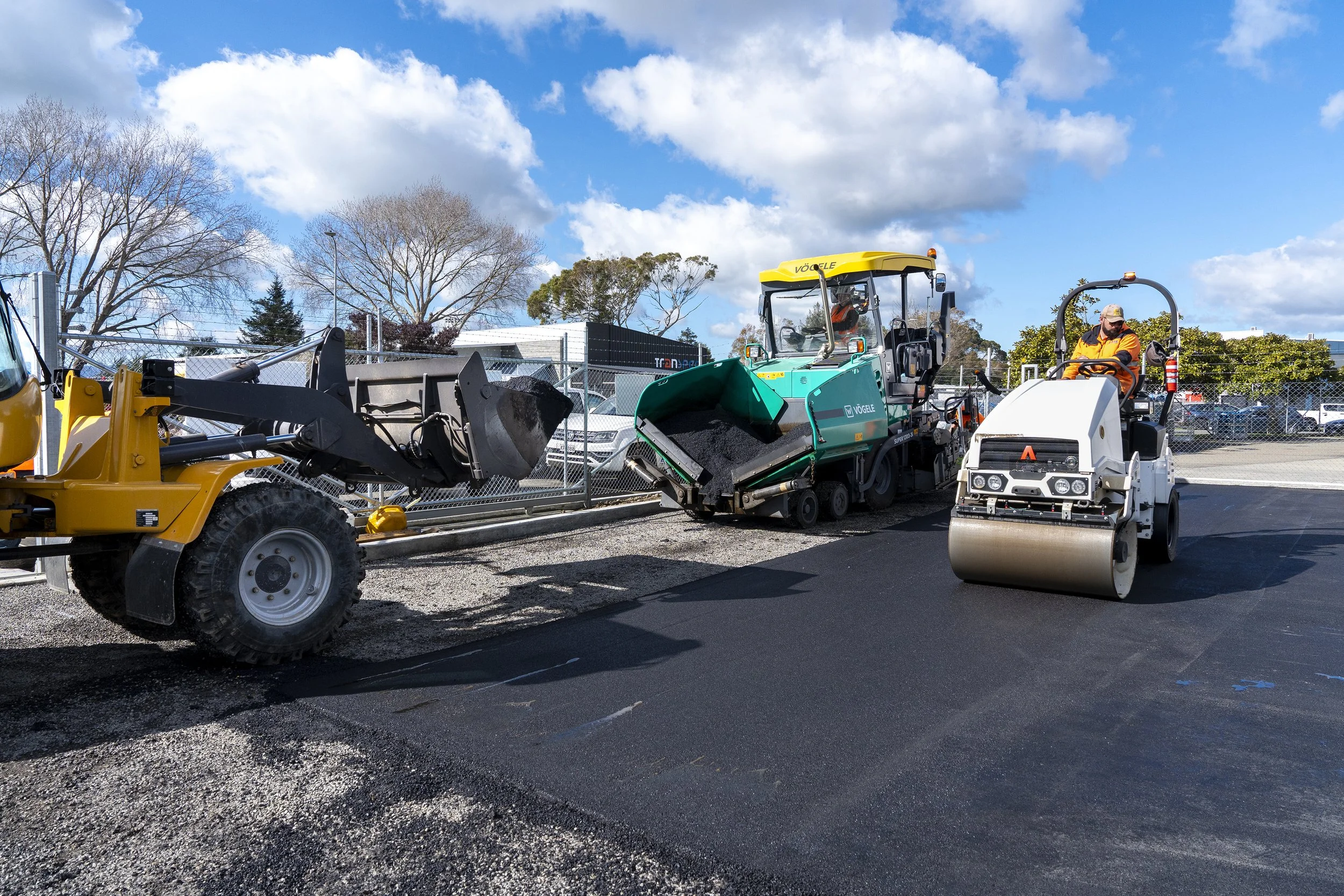 Workers paving a road with asphalt using road roller and asphalt paver machine under a partly cloudy sky