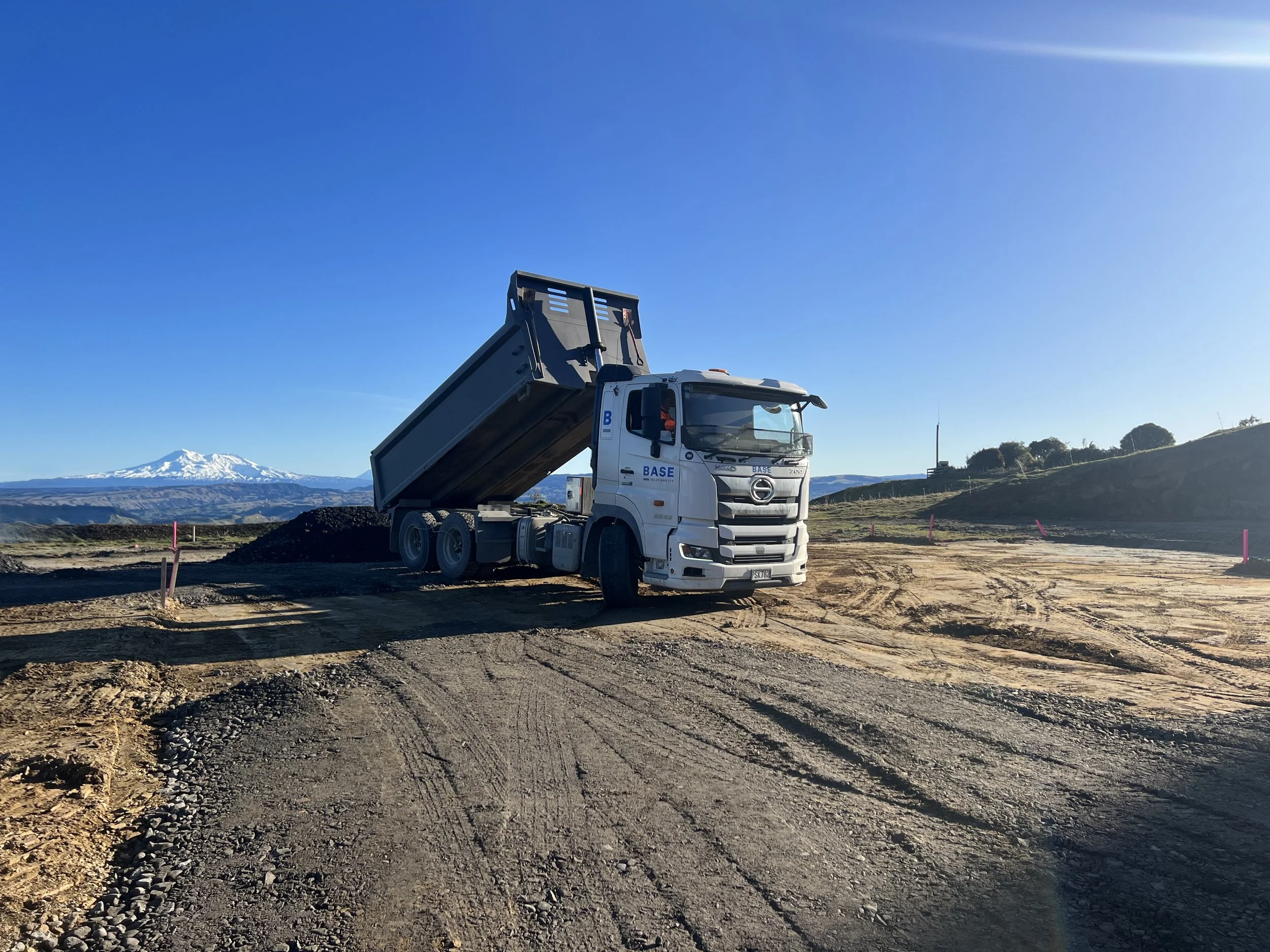 A white dump truck with its bed raised, parked on a dirt construction site with gravel, with snow-capped mountains in the background under a clear blue sky.