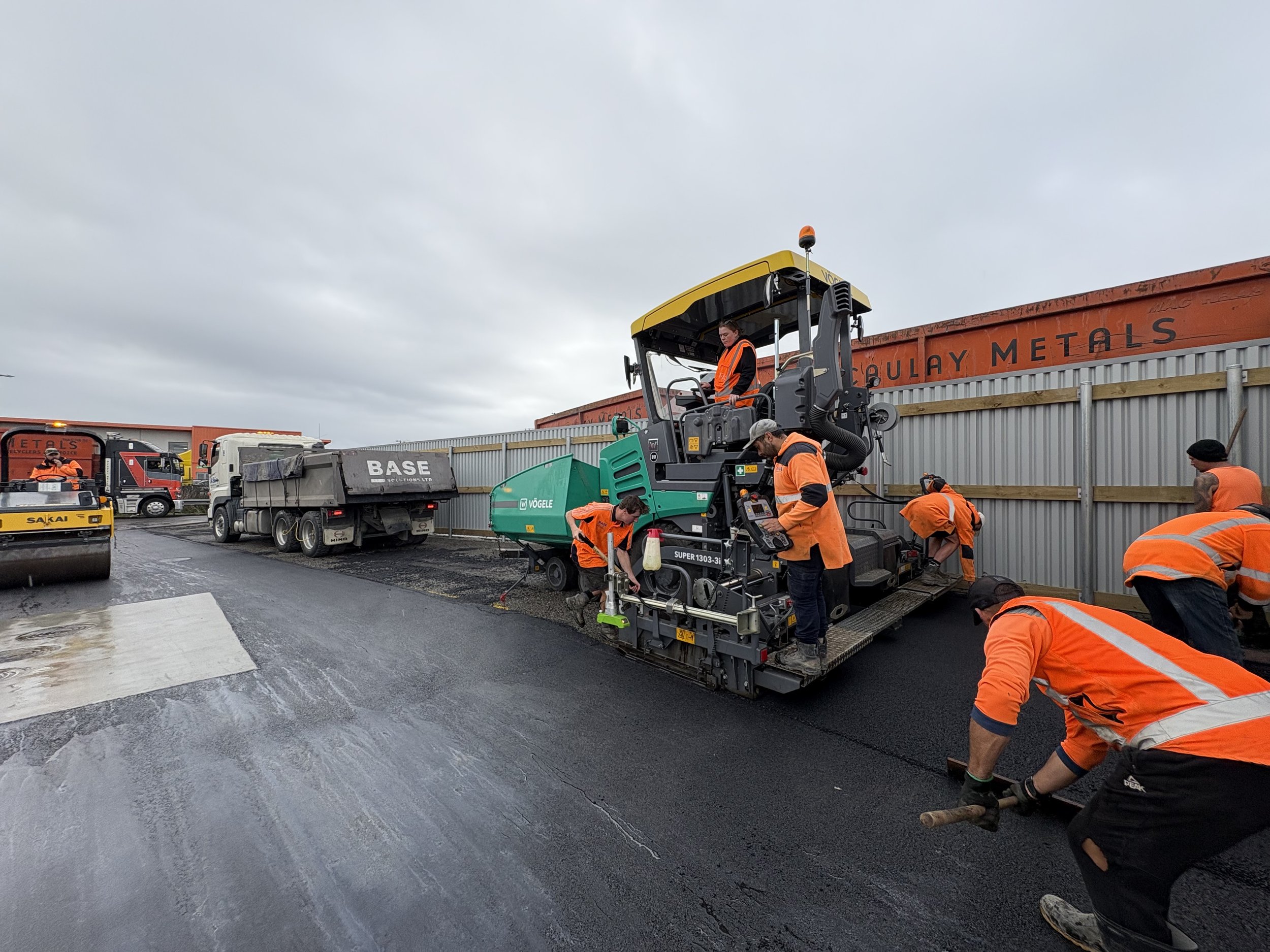 Construction workers paving a road with asphalt using heavy machinery, with trucks and a metal fence in the background under a cloudy sky.