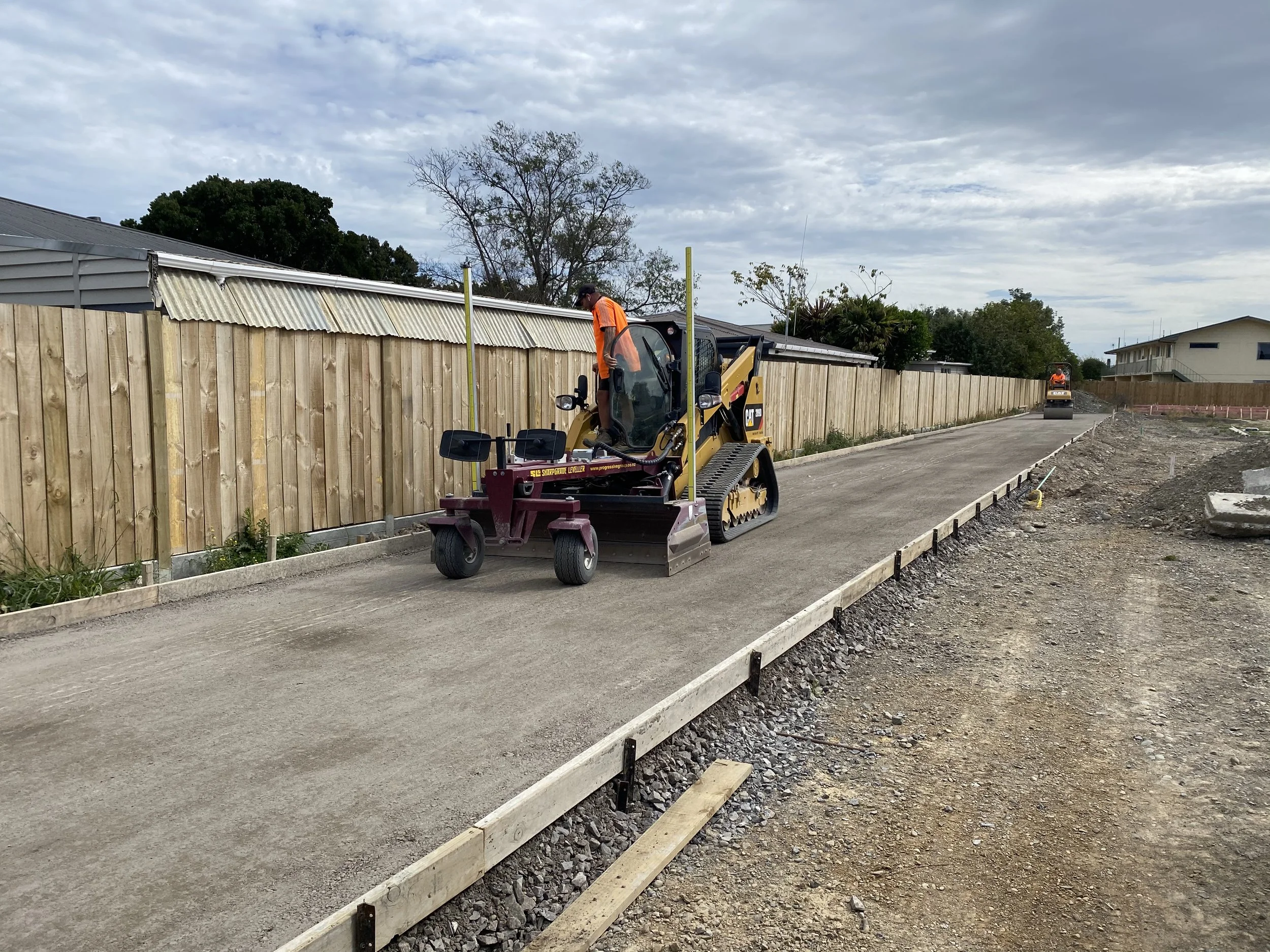 Workers using a small construction roller to pave a new road in a suburban area, with a wooden fence and houses in the background.