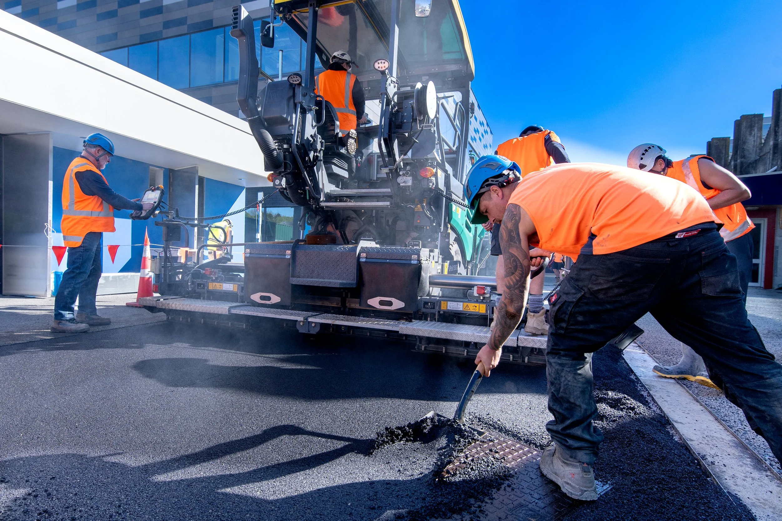 Workers in orange vests and hard hats paving a road with asphalt using a heated asphalt paver machine under a clear blue sky.