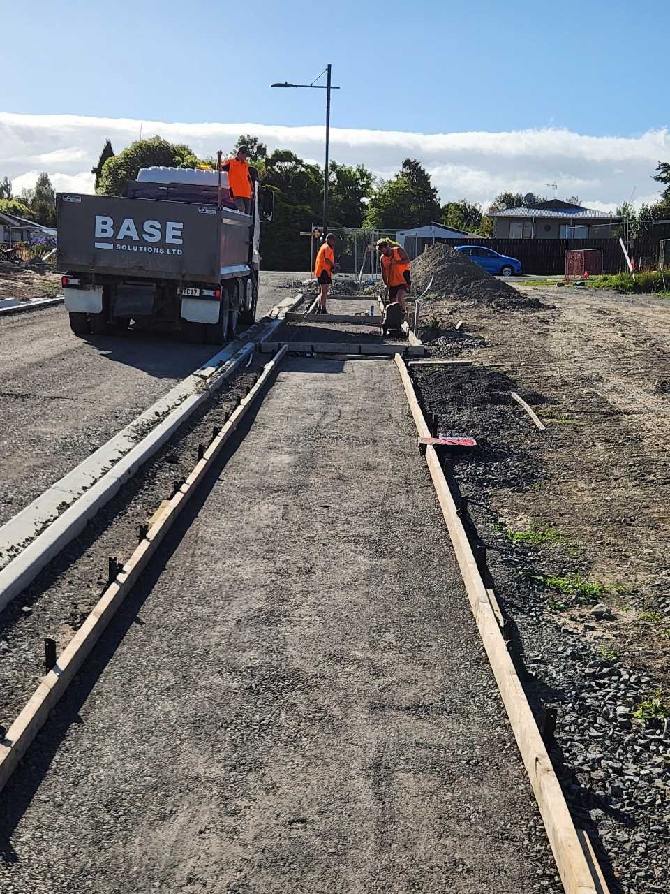 Construction workers laying down new pavement on a sidewalk or pathway, with wooden forms in place and a truck nearby.