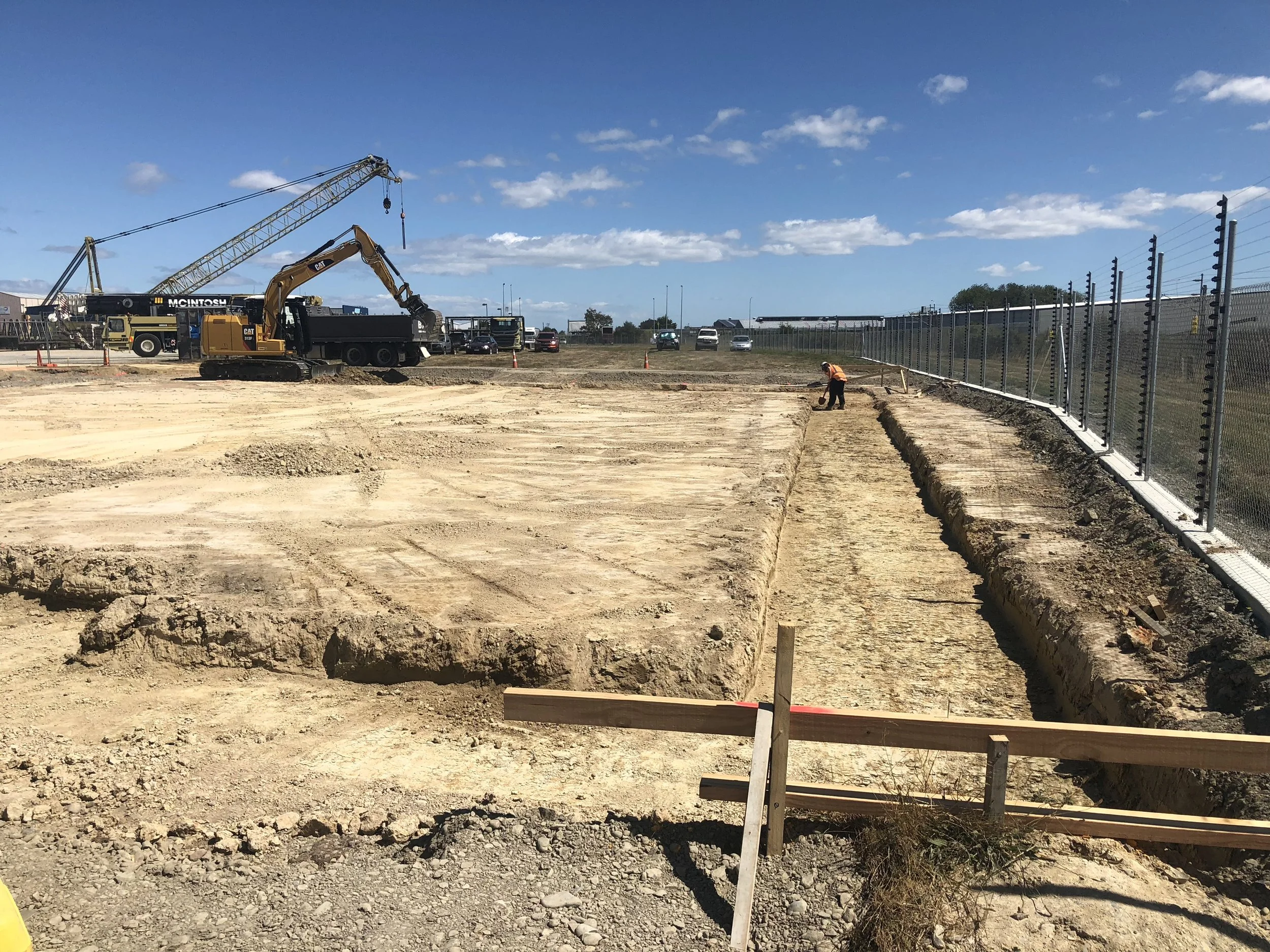 Construction site with excavated dirt, a worker, heavy machinery, and a fence along the right side, with parked cars and a crane in the background under a blue sky with clouds.