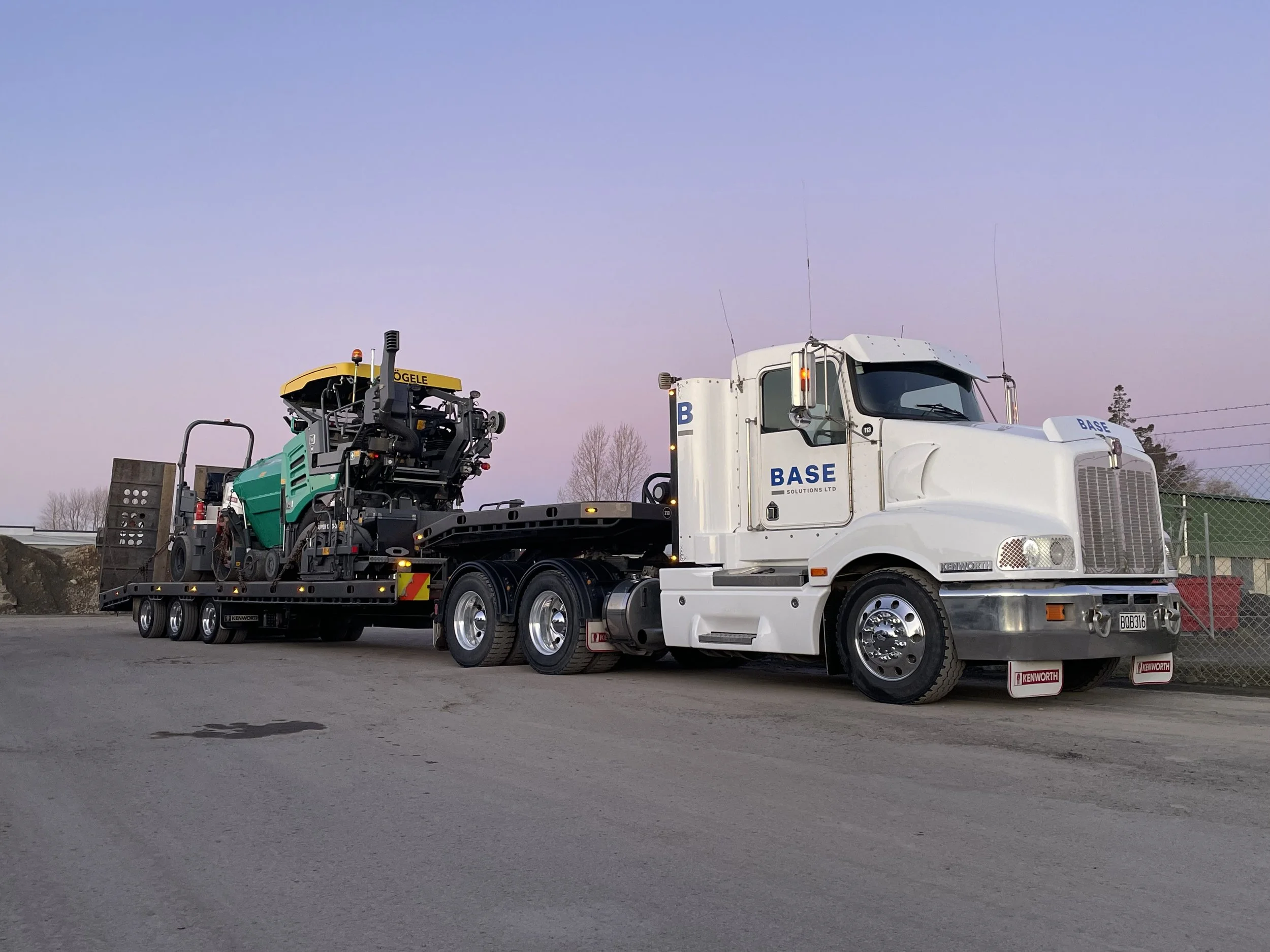 A white semi-truck is carrying construction equipment on a flatbed trailer, parked outdoors during dusk with a pink and blue sky.