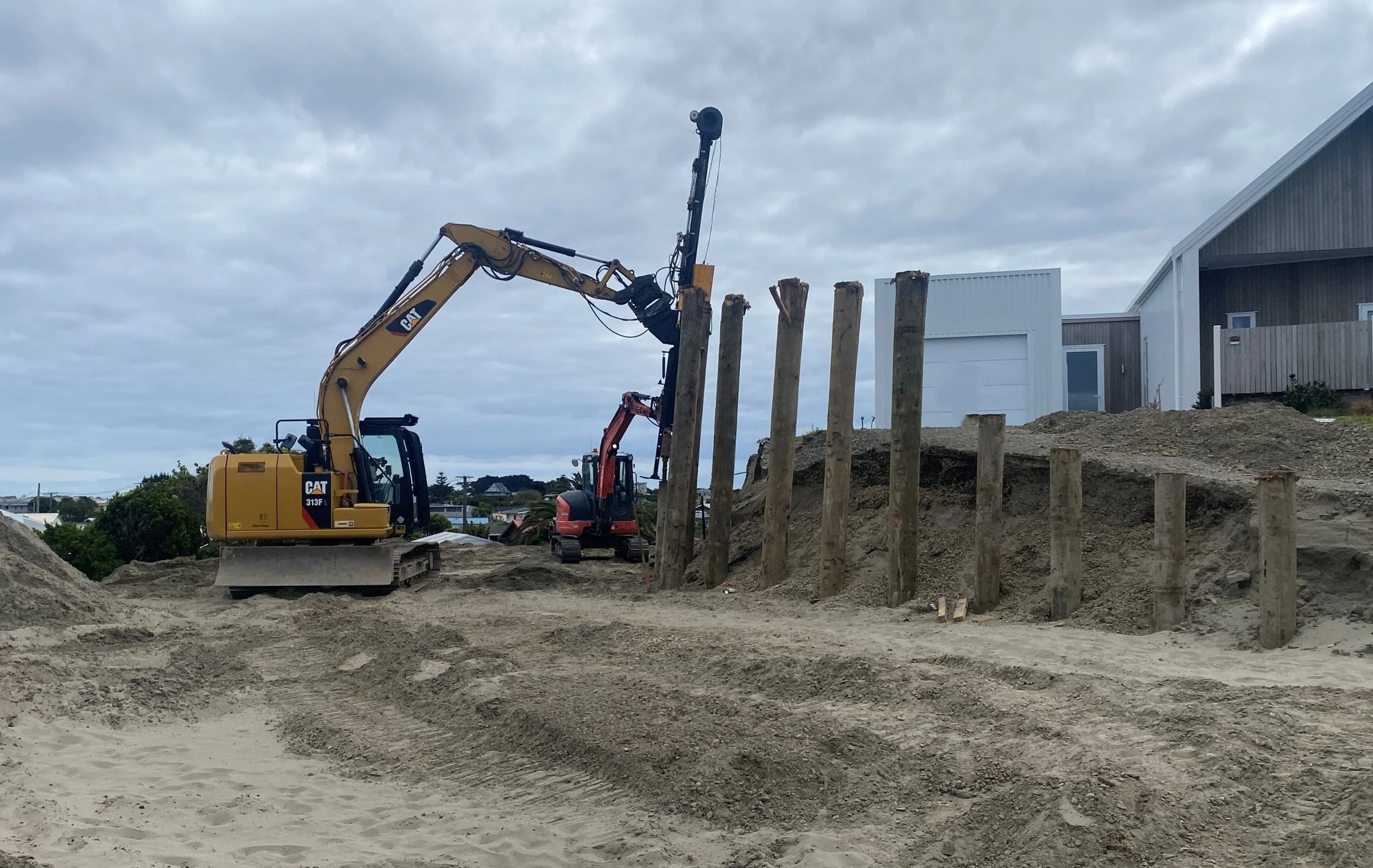 Construction site with a yellow excavator and a smaller pink excavator, wooden pillars, dirt, and residential buildings in the background under a cloudy sky.
