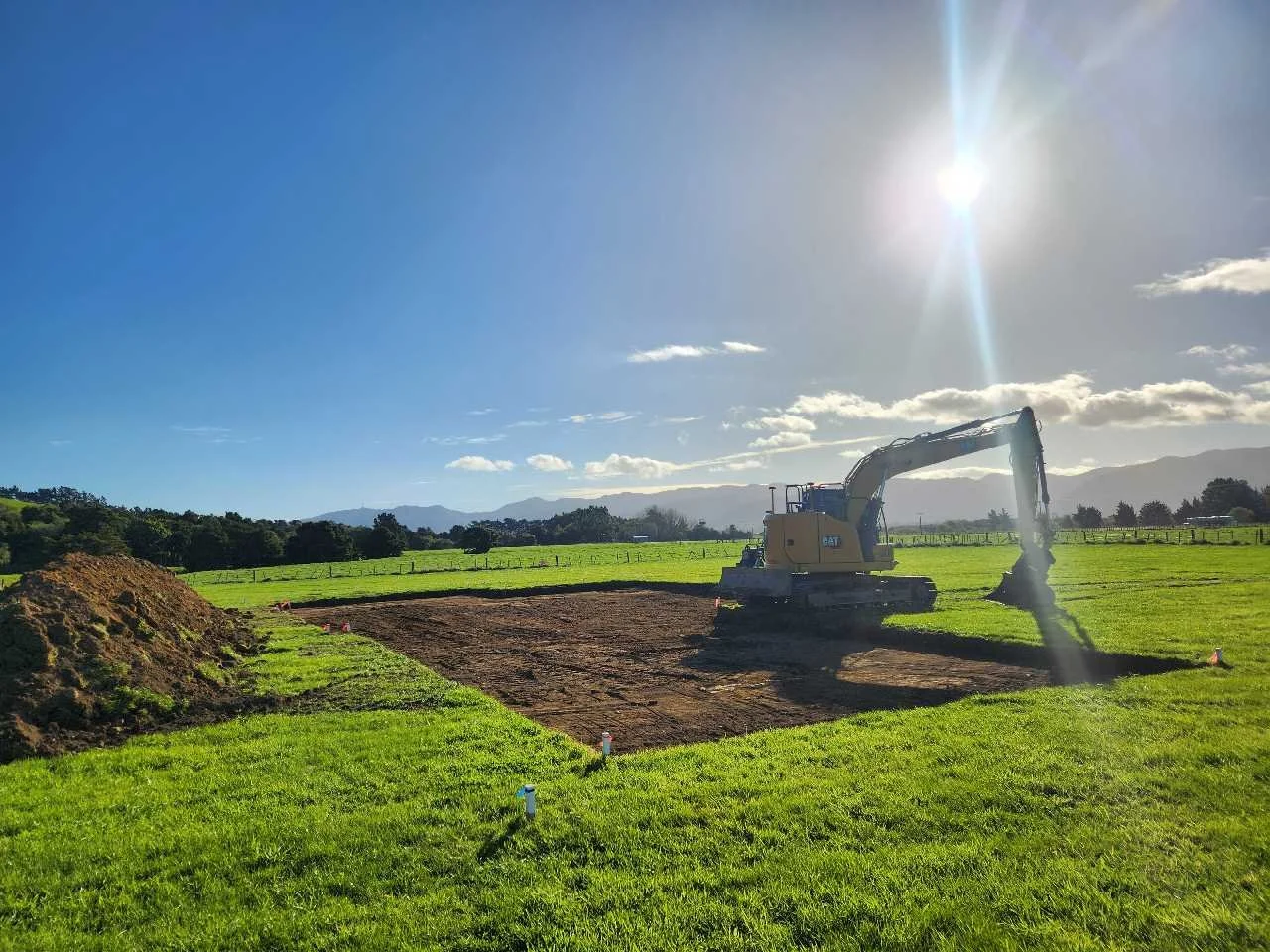Construction site in a grassy field with a yellow excavator moving soil, under a blue sky with the sun shining and some clouds.