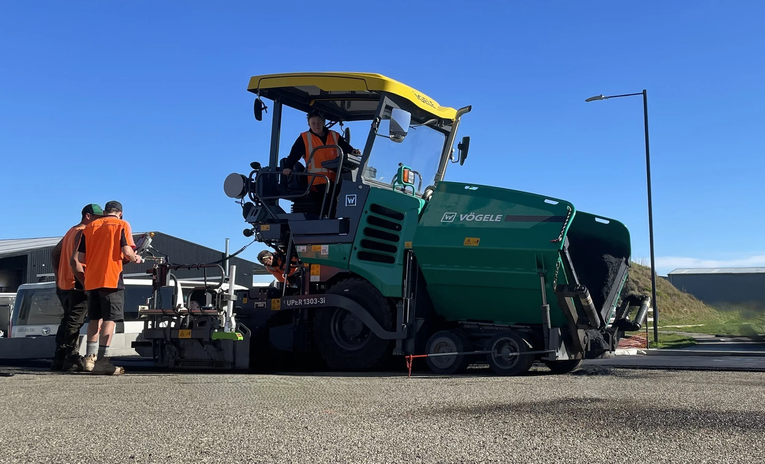 Construction workers in orange vests operating a large green Vögele asphalt paving machine on a road, with a clear blue sky above and industrial buildings in the background.