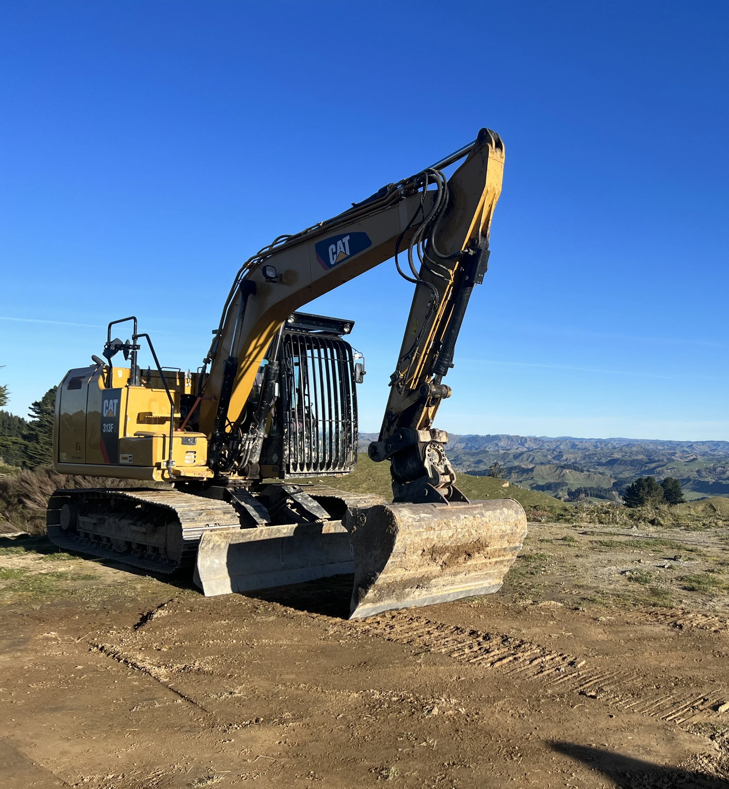 A yellow Caterpillar excavator on a dirt construction site with mountains in the background and clear blue sky.