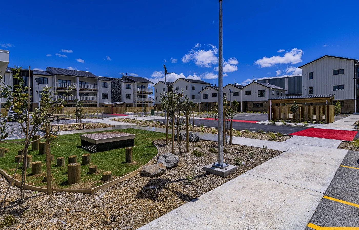 Modern residential neighborhood with apartment buildings, a small park area with trees, a bench, and walking paths under a partly cloudy sky.