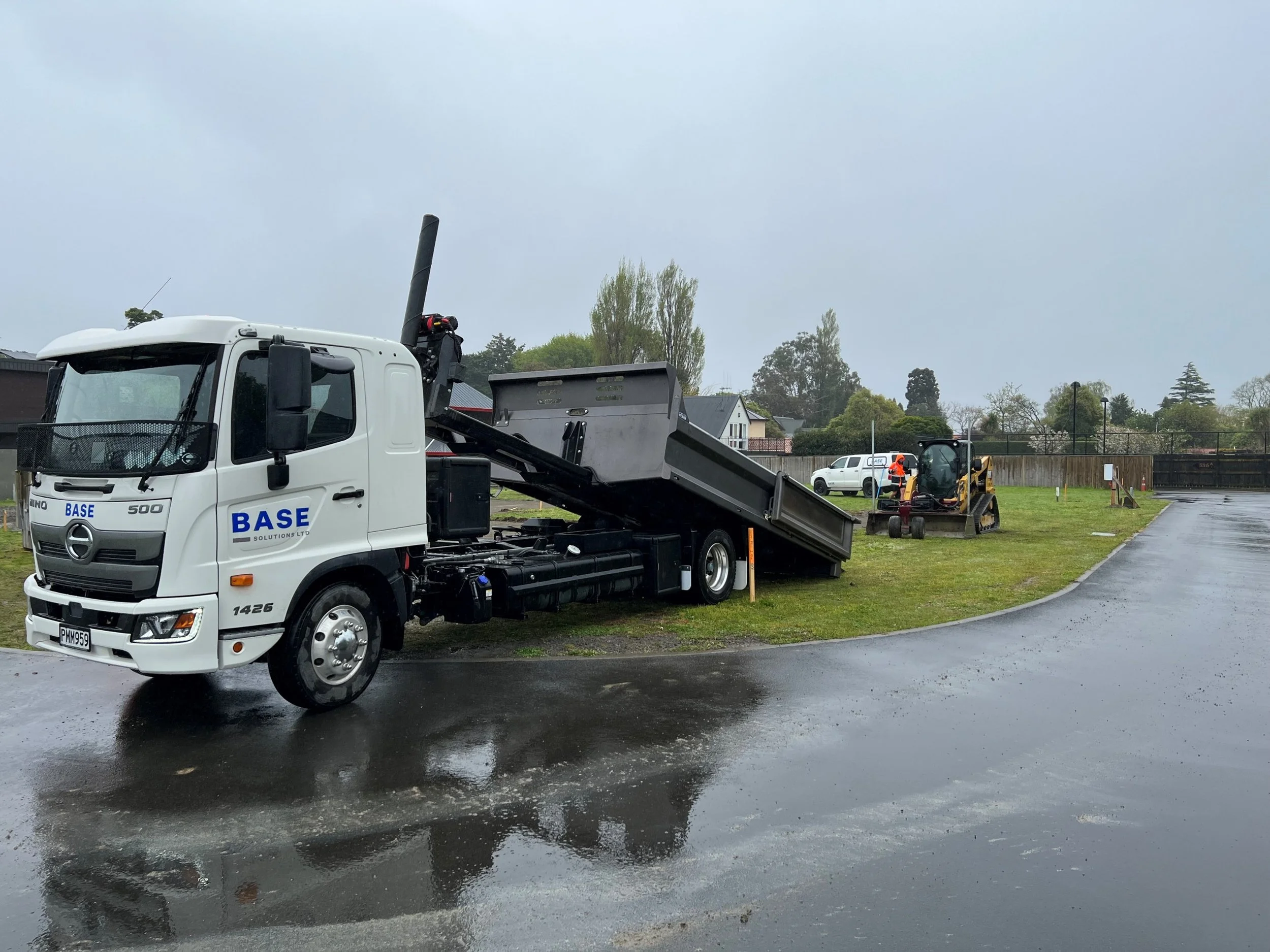 A white flatbed truck with the logo 'BASE' on the side, parked on a wet road with construction equipment and workers in the background.